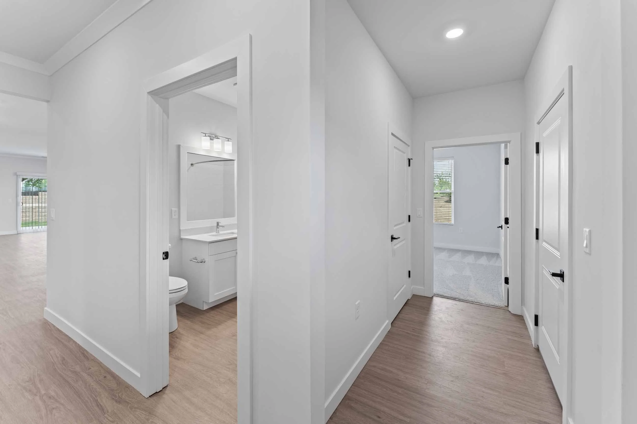 Interior view of a modern house with white walls and doors, showing a hallway leading to a room with a window, and a small bathroom with a mirror and sink visible on the left.