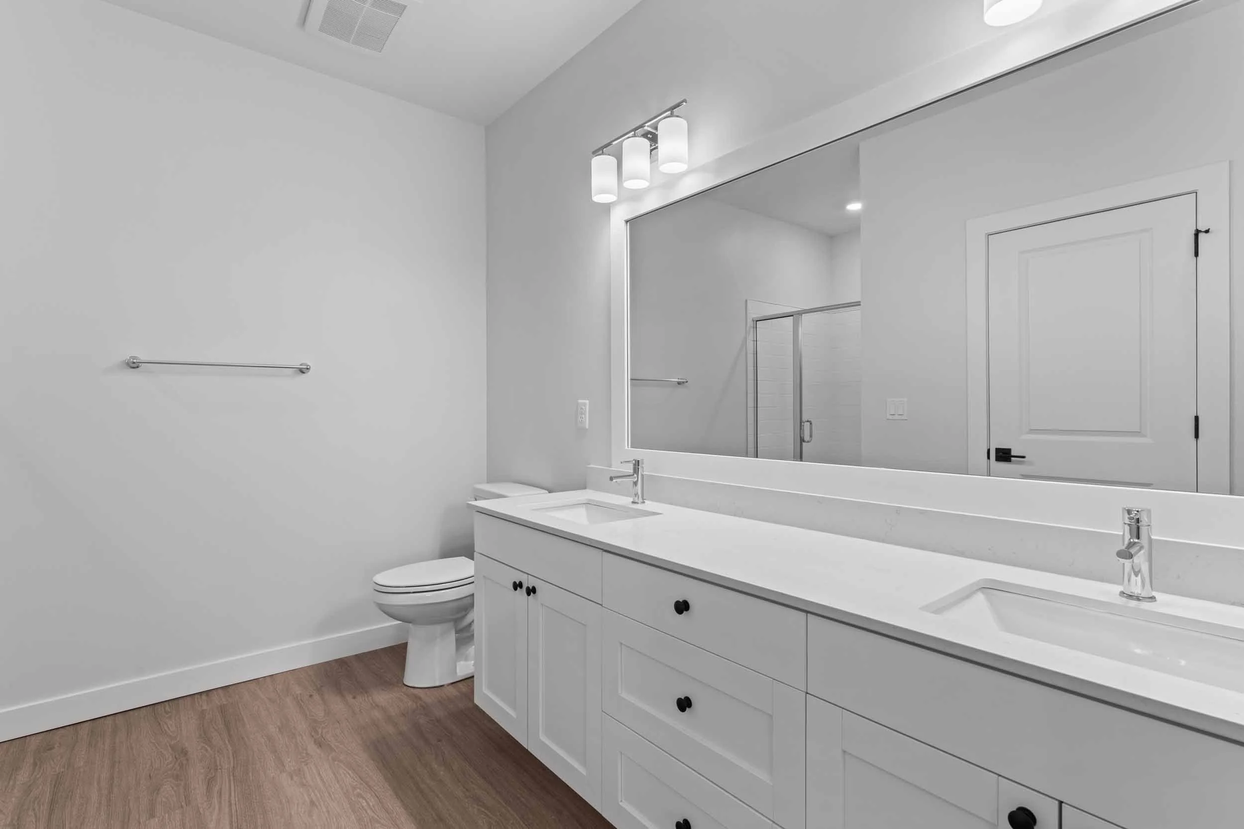 A modern bathroom with white walls, a white vanity with black knobs, a large mirror, a white toilet, a shower with glass door, and wood flooring. There is a three-light fixture above the mirror and a towel bar on the wall.