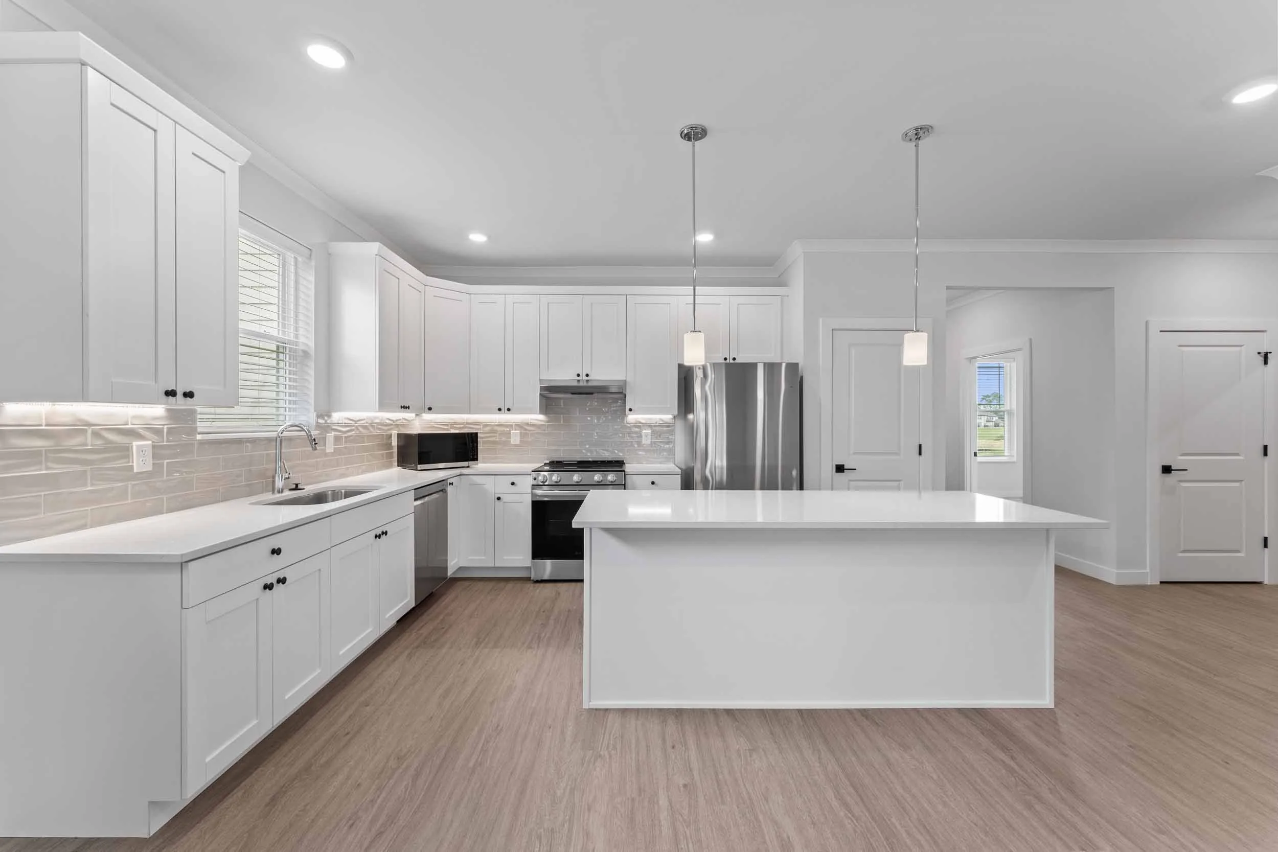 Modern white kitchen with island, white cabinets, stainless steel appliances, pendant lights, and wood flooring.