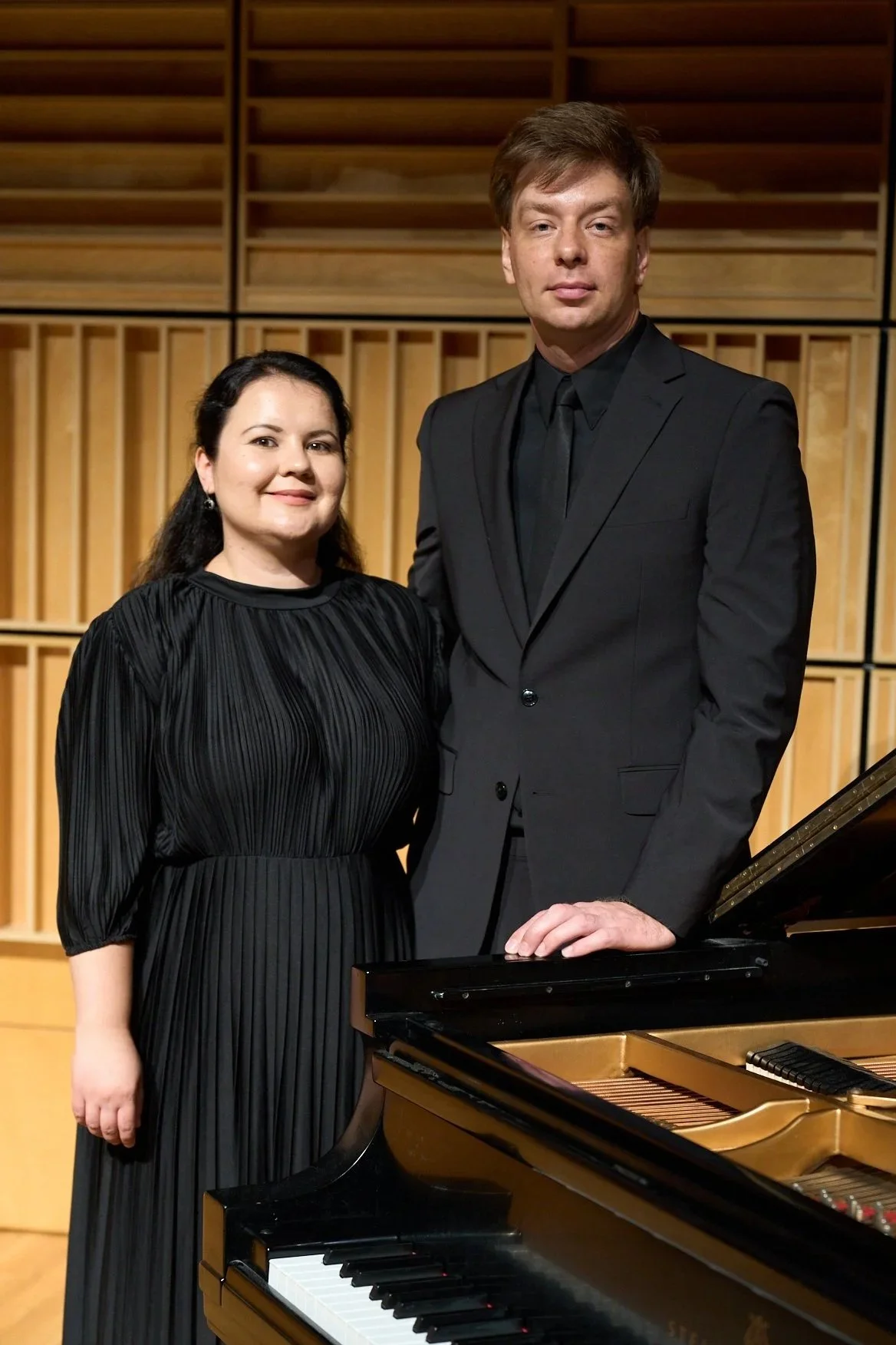 The Skobin Duo (Diana Skobina and Sergei Skobin) in all-black concert attire, posing next to a grand piano in a concert hall.