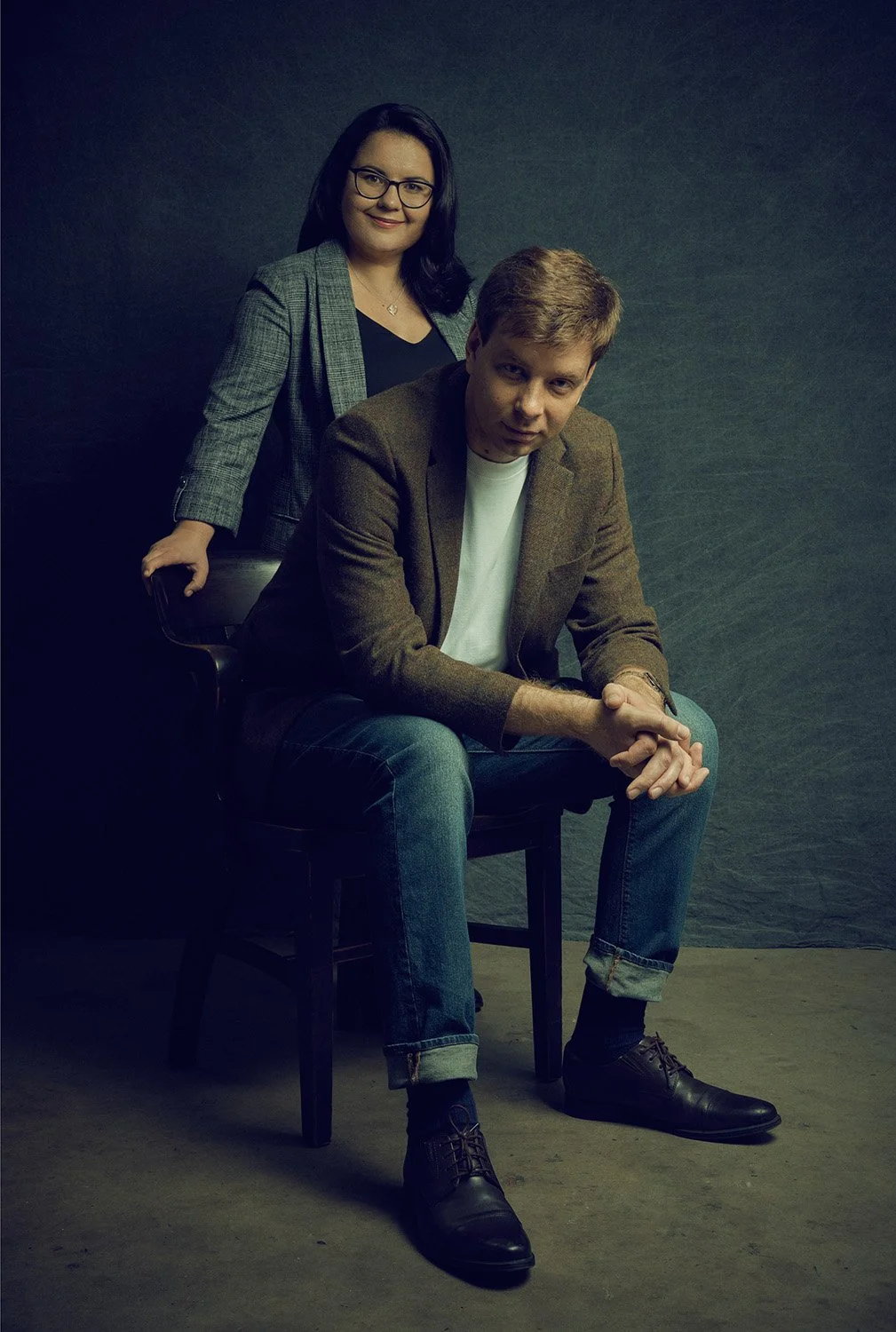Studio portrait of classical piano duo Skobin Duo. Sergei Skobin is seated on a chair while Diana Skobina stands behind him against a green and black backdrop, gazing forward.