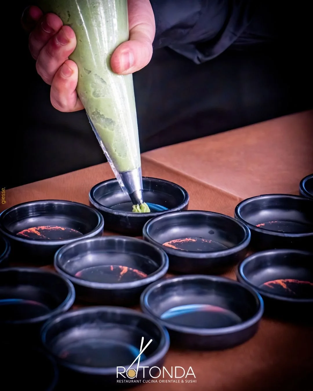 A person filling small black dishes with green wasabi using a piping bag at a restaurant. The table has multiple dishes and there is a logo at the bottom that says 'Rotonda - Restaurant Cucina Orientale & Sushi'.