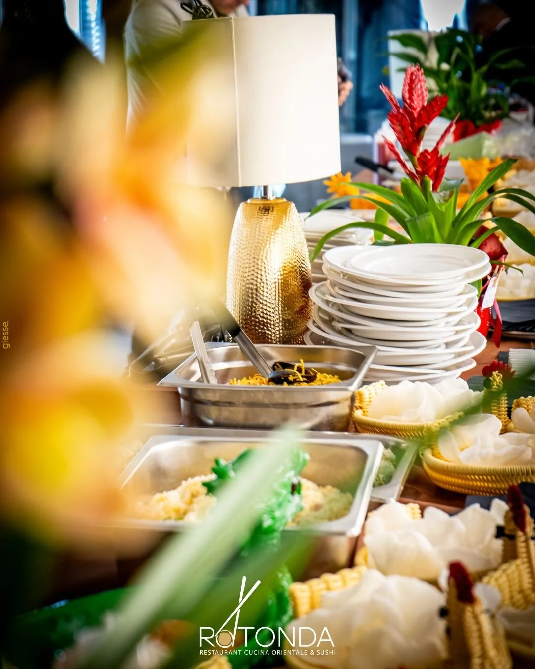 Tavolo con piatti bianchi impilati, lampada dorata, piante verdi e fiori rossi, alimenti e piatti pronti in buffet in un ristorante orientale.