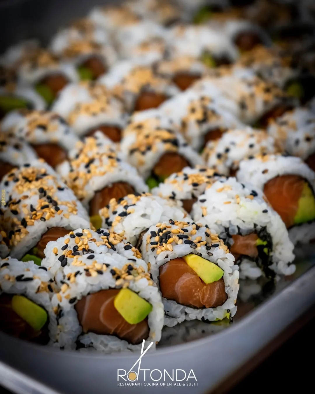 Riunione di sushi con salmone e avocado, coperto di semi di sesamo, in un piatto di ceramica. Logo di Rotorna, ristorante di cucina orientale e sushi.