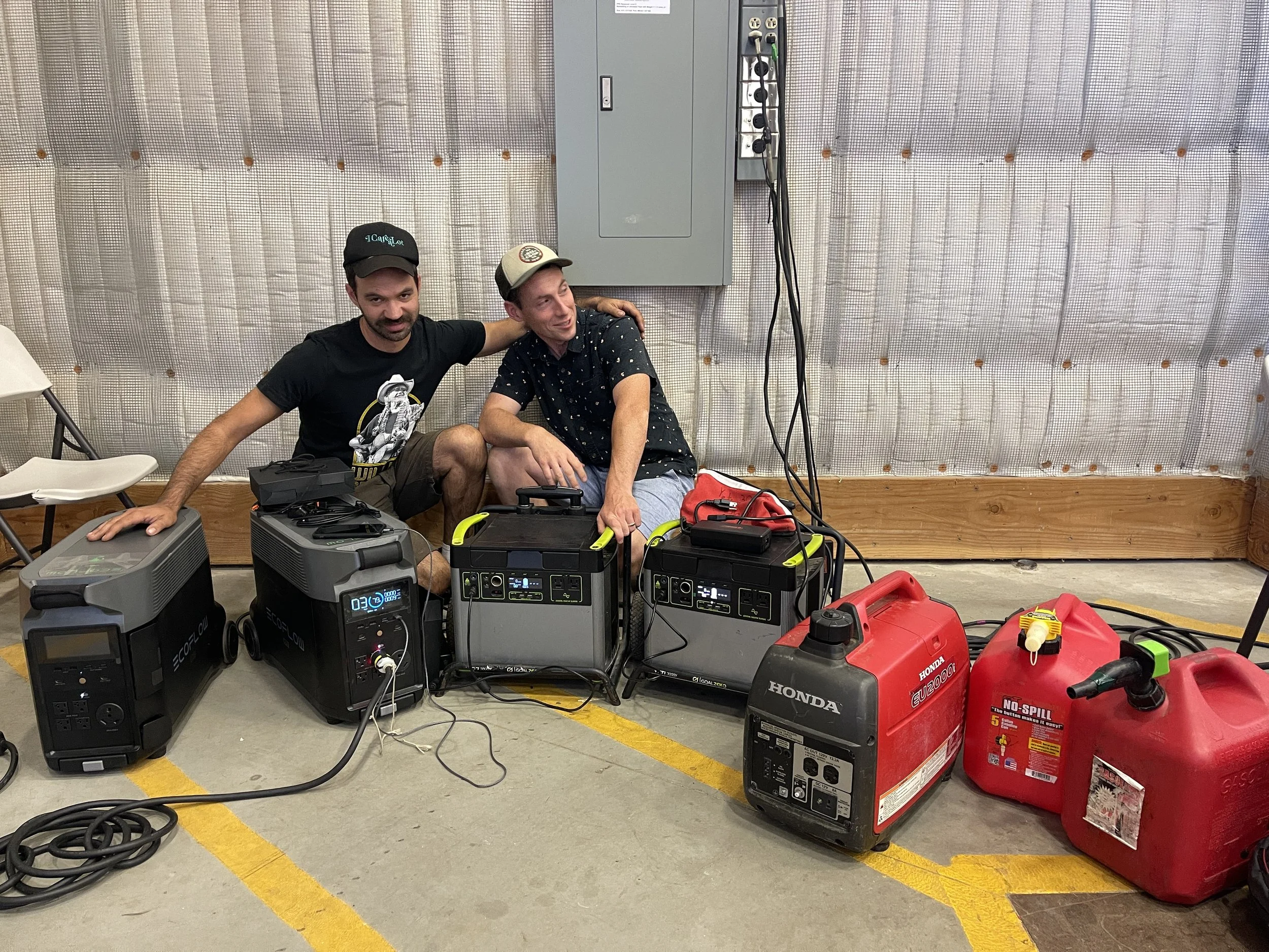 Two men sitting on the floor among equipment including generators, fuel containers, and cables, in a room with a plastic-covered wall behind them.