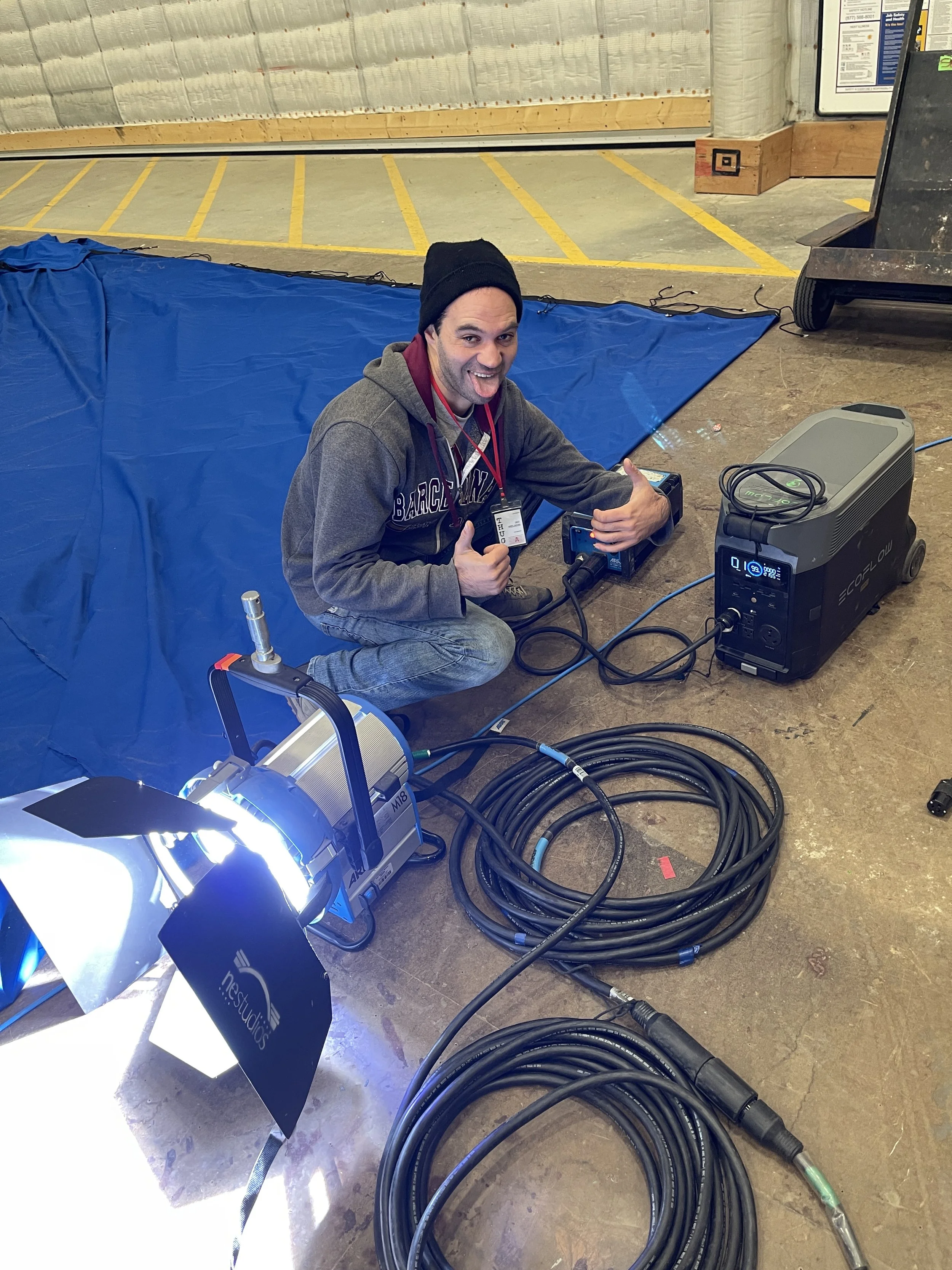 A man kneeling on the floor, smiling and giving a thumbs up, surrounded by filming or lighting equipment, including a large light, cables, and a generator, indoors with a blue backdrop and parking lot lines visible in the background.