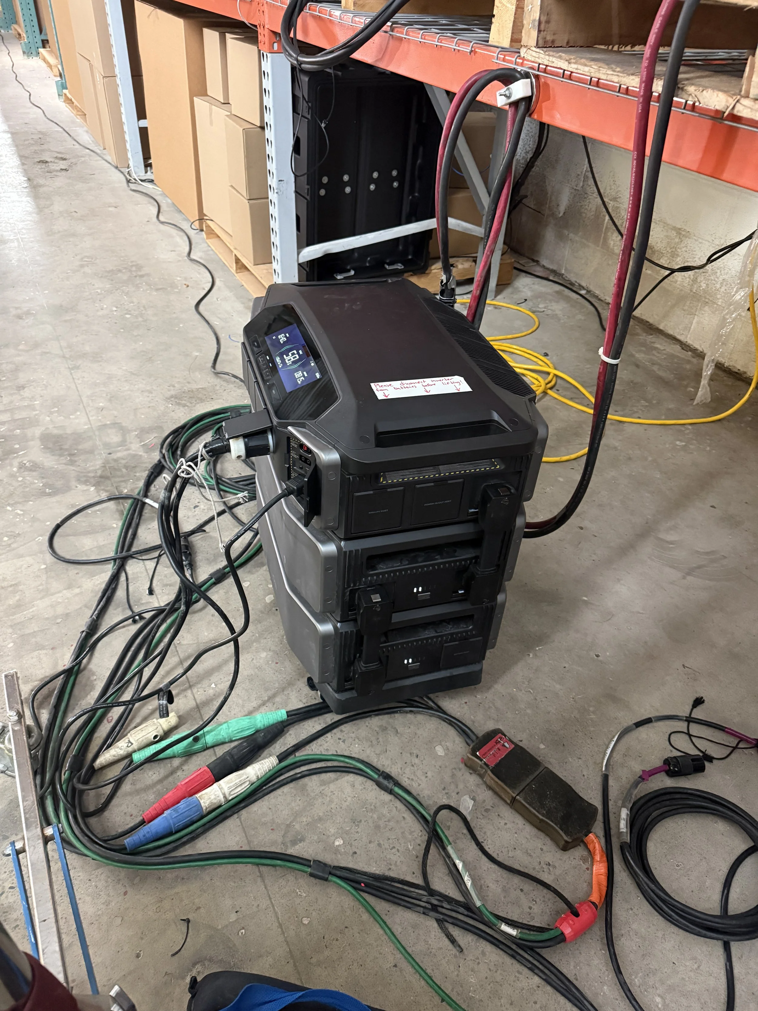 A black portable generator with various cables and equipment connected, placed on a concrete floor in a warehouse setting with shelves and boxes in the background.