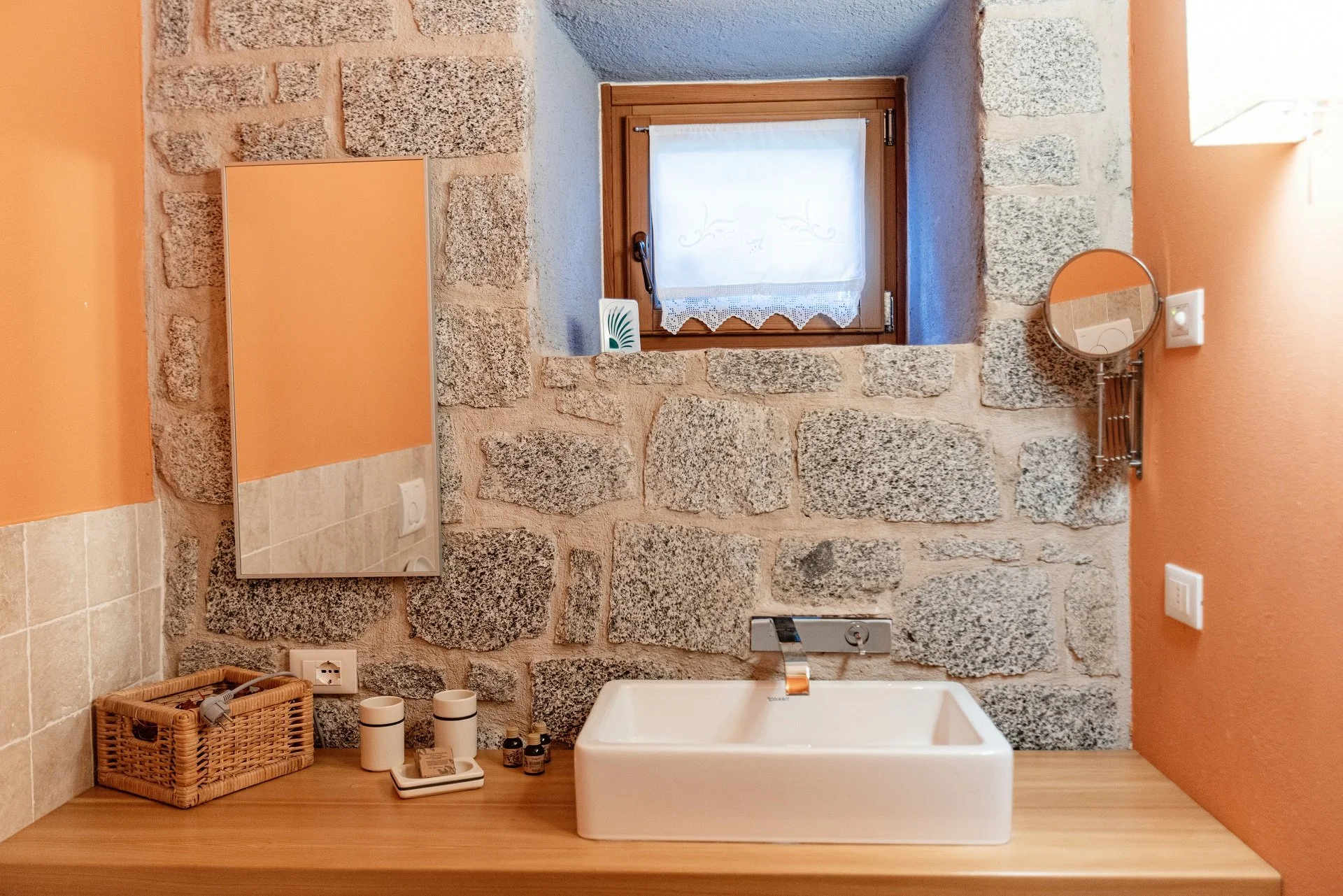 Un bagno con pareti di pietra, un piccolo finestrino con tende in pizzo, un mobile con ripiano in legno, un lavabo bianco rettangolare, uno specchio rotondo, alcuni barattoli e un cesto di vimini sulla superficie misma.