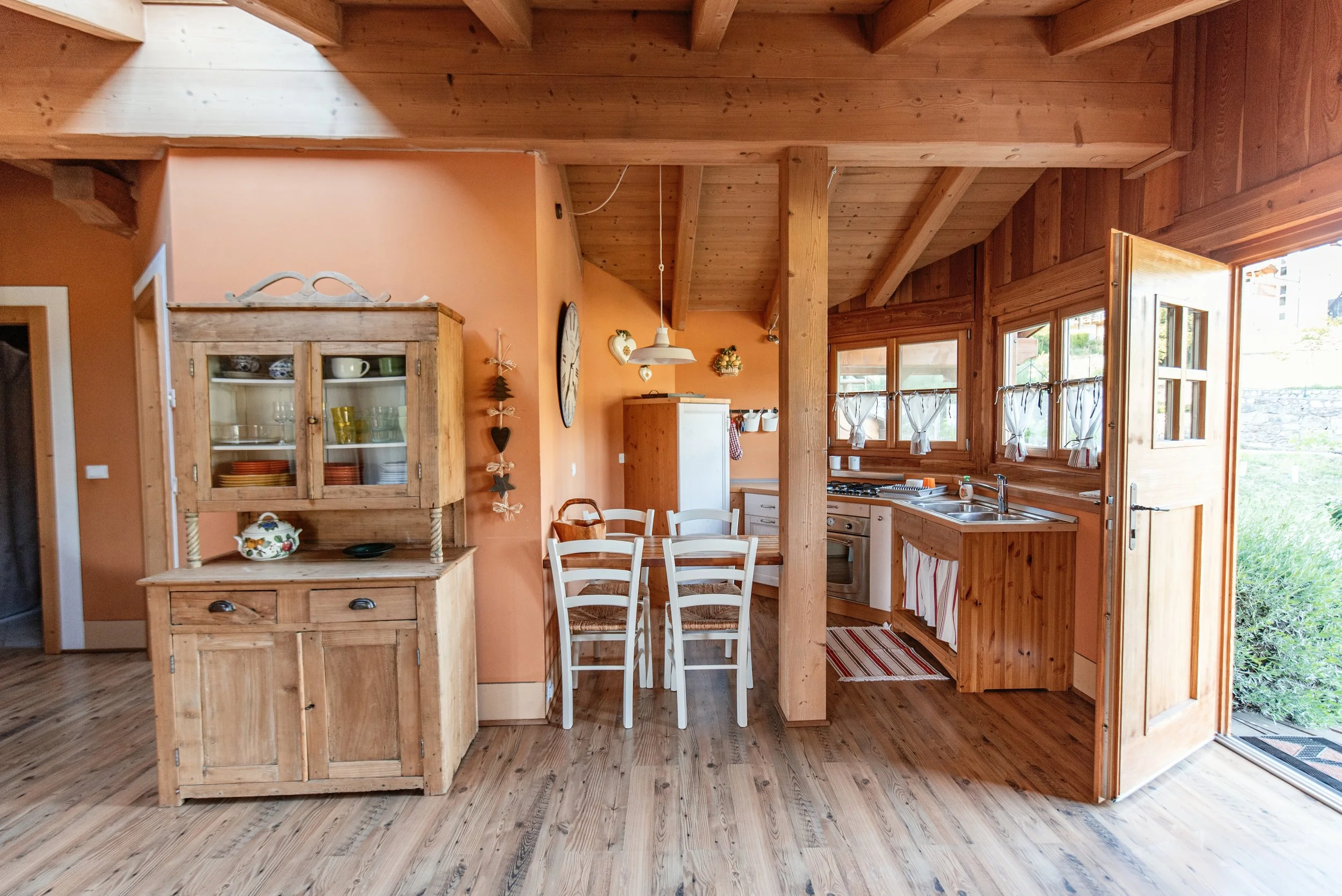 Cucina rustica in legno con tavolo e sedie bianche, mobili in legno, finestre con tende a righe rosse e bianche, pavimento in legno, pareti color terracotta, soffitto con travi in legno.