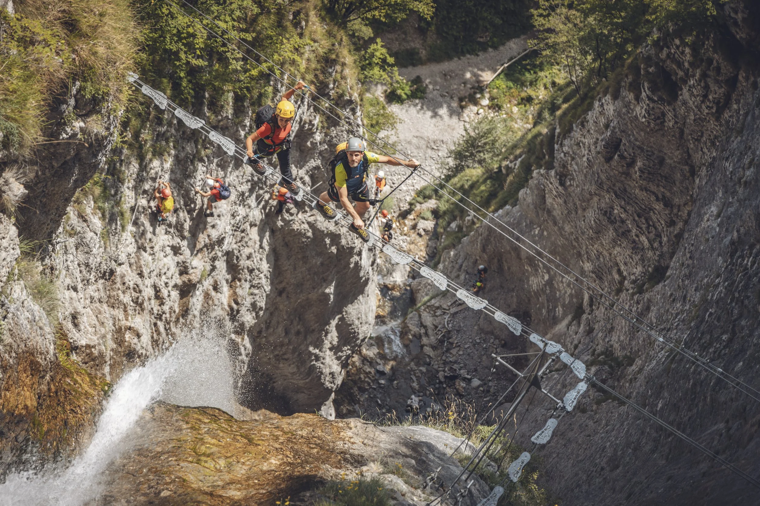 Escursionisti che attraversano un tratto di corda sopra una cascata in una stretta forra rocciosa.