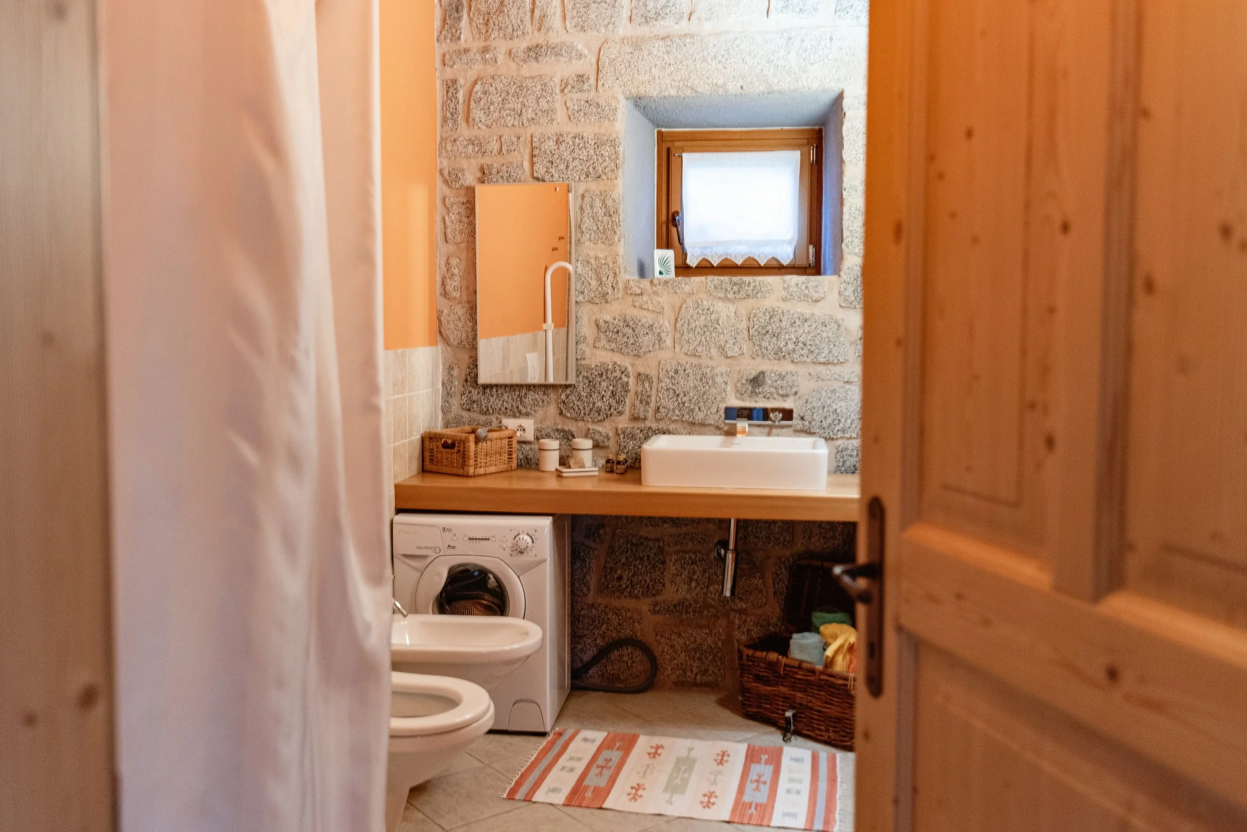 Vista di un bagno rustico con pareti di pietra, un lavandino bianco su un piano di legno, una lavatrice, un piccolo finestrino e un tappeto colorato sul pavimento.