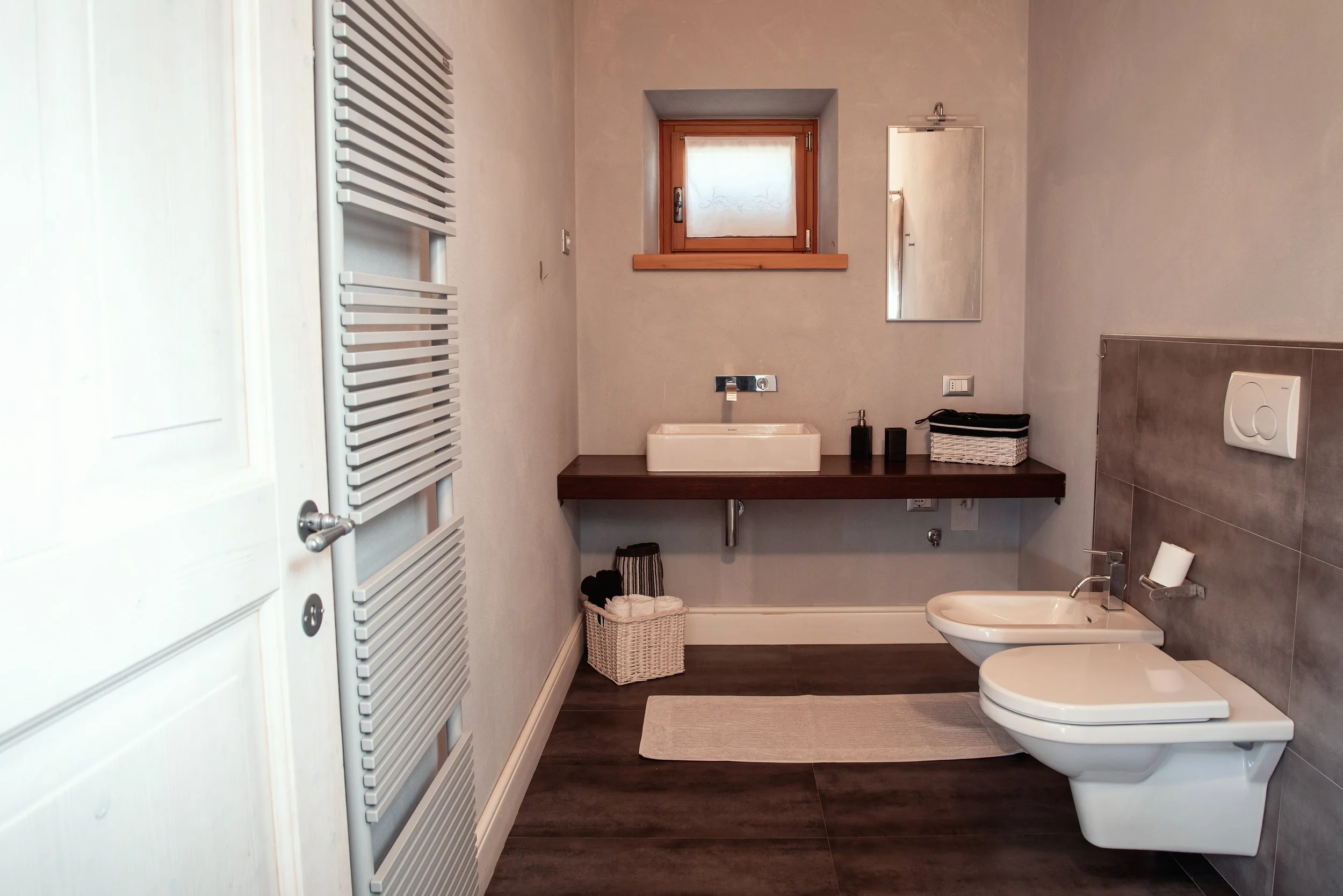Bagno moderno con sanitari bianchi, lavabo su piano di legno, finestra in alto, specchio, e asciugamani.