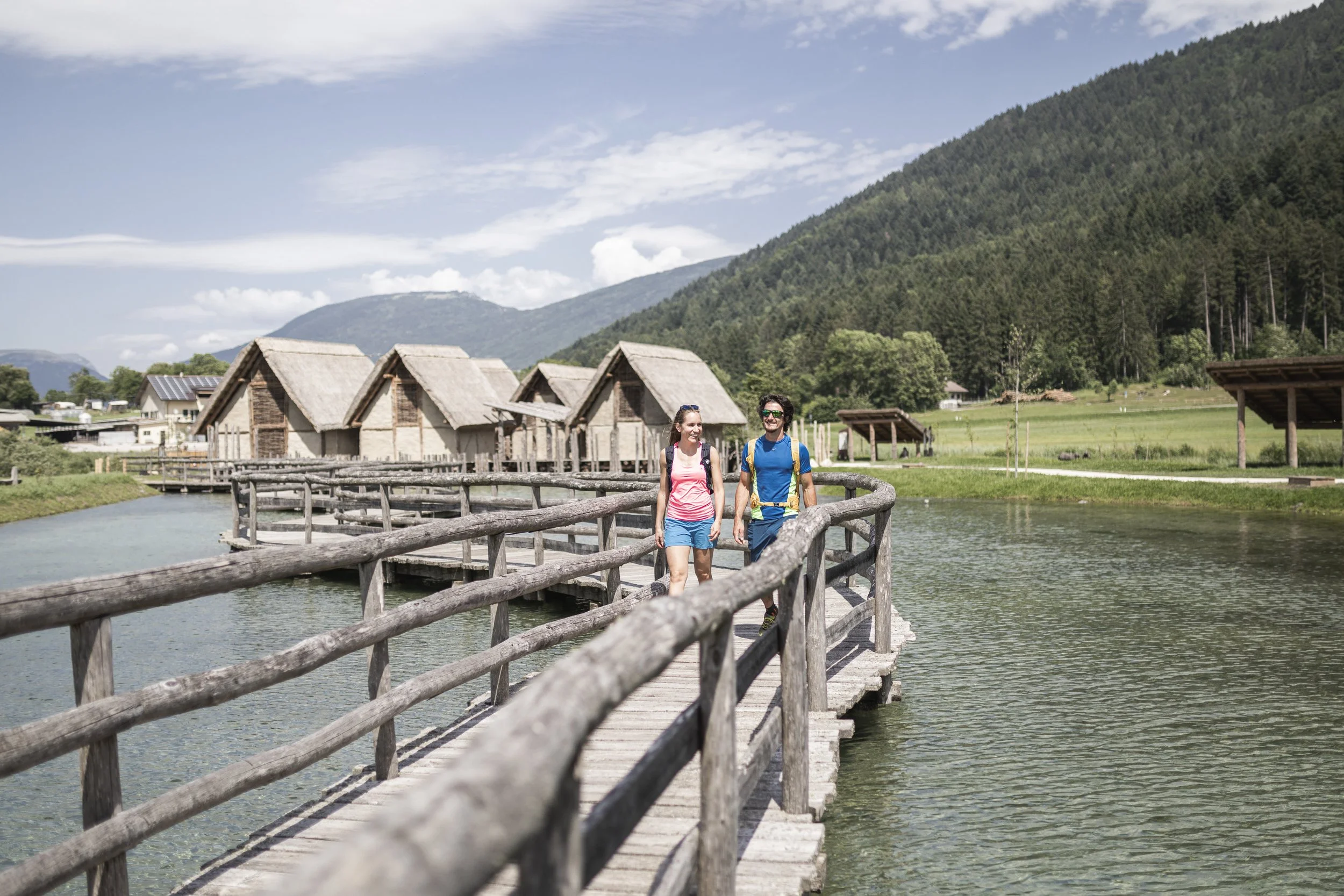 Due persone che camminano sul ponte di legno sopra un lago in un paesaggio naturale con capanne di legno, montagne e cielo nuvoloso sullo sfondo.