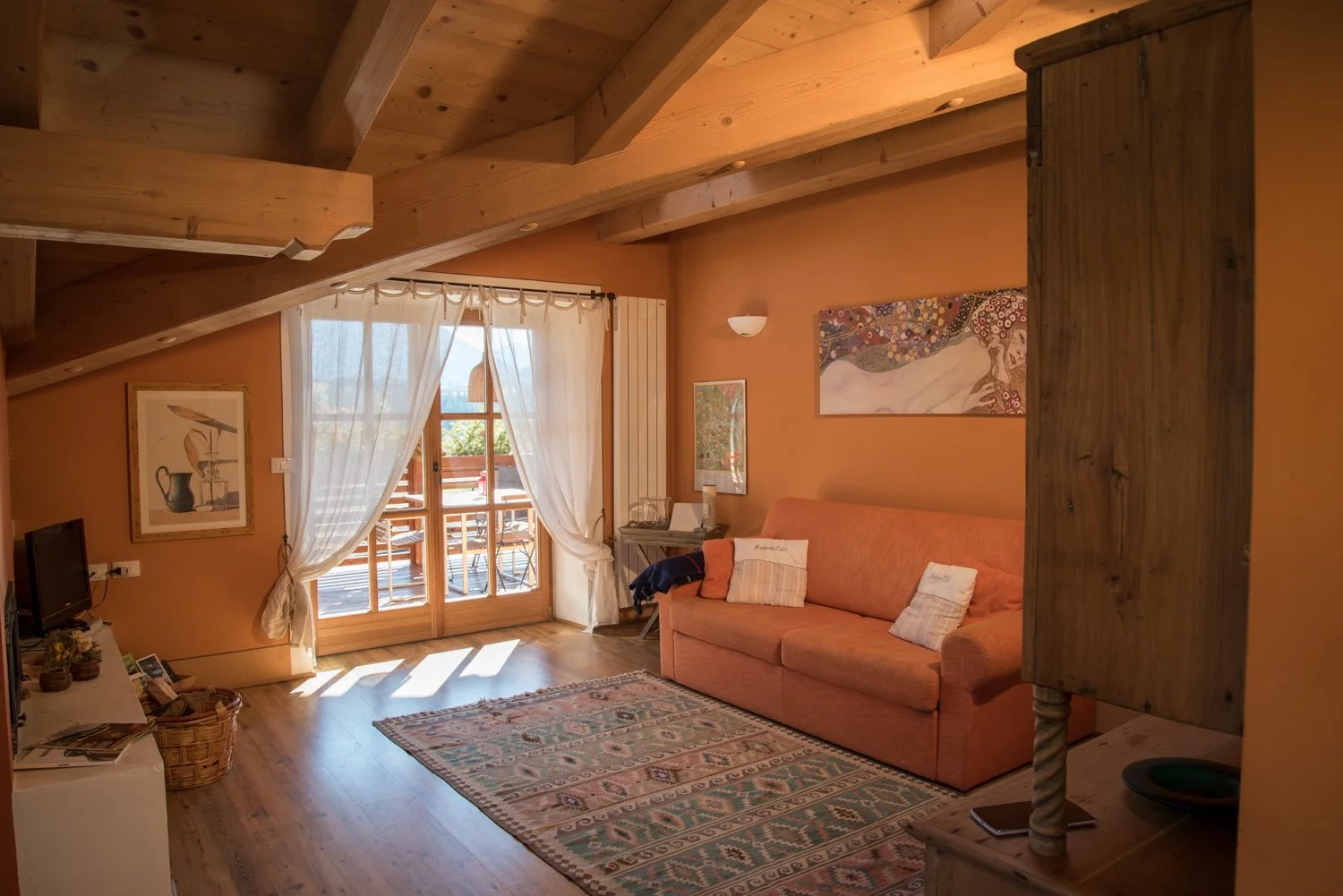 Interno di un soggiorno con pareti arancioni, divano rosa, tappeto e pareti decorate con quadri. Porta finestra che si apre su un balcone con sedie e tavolo di legno, tende bianche leggere. Sopra ci sono travi di legno a vista.