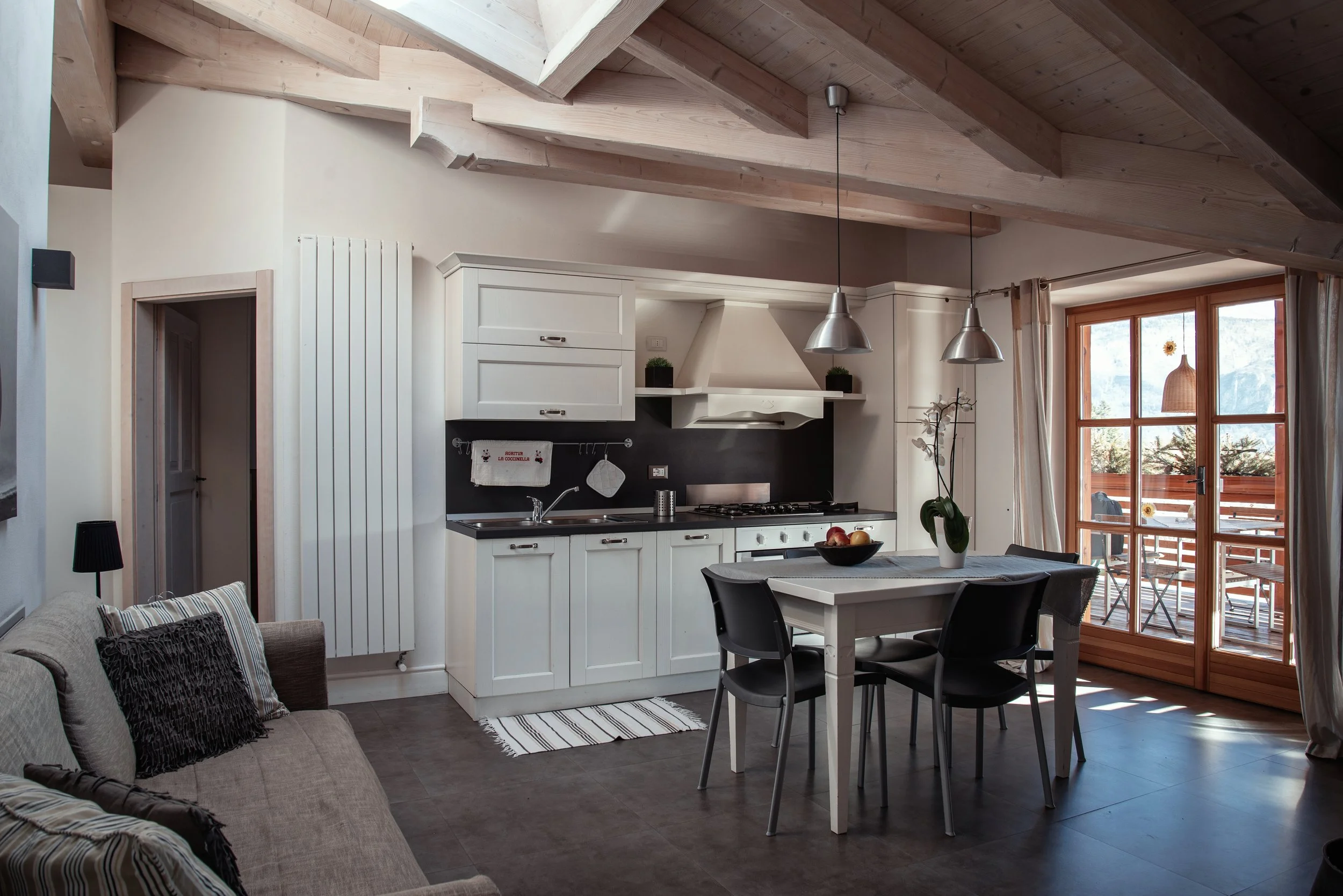 Interno di una cucina con pareti bianche, soffitto in legno, tavolo bianco con sedie nere, soggiorno con divano e un grande balcone con vista esterna.