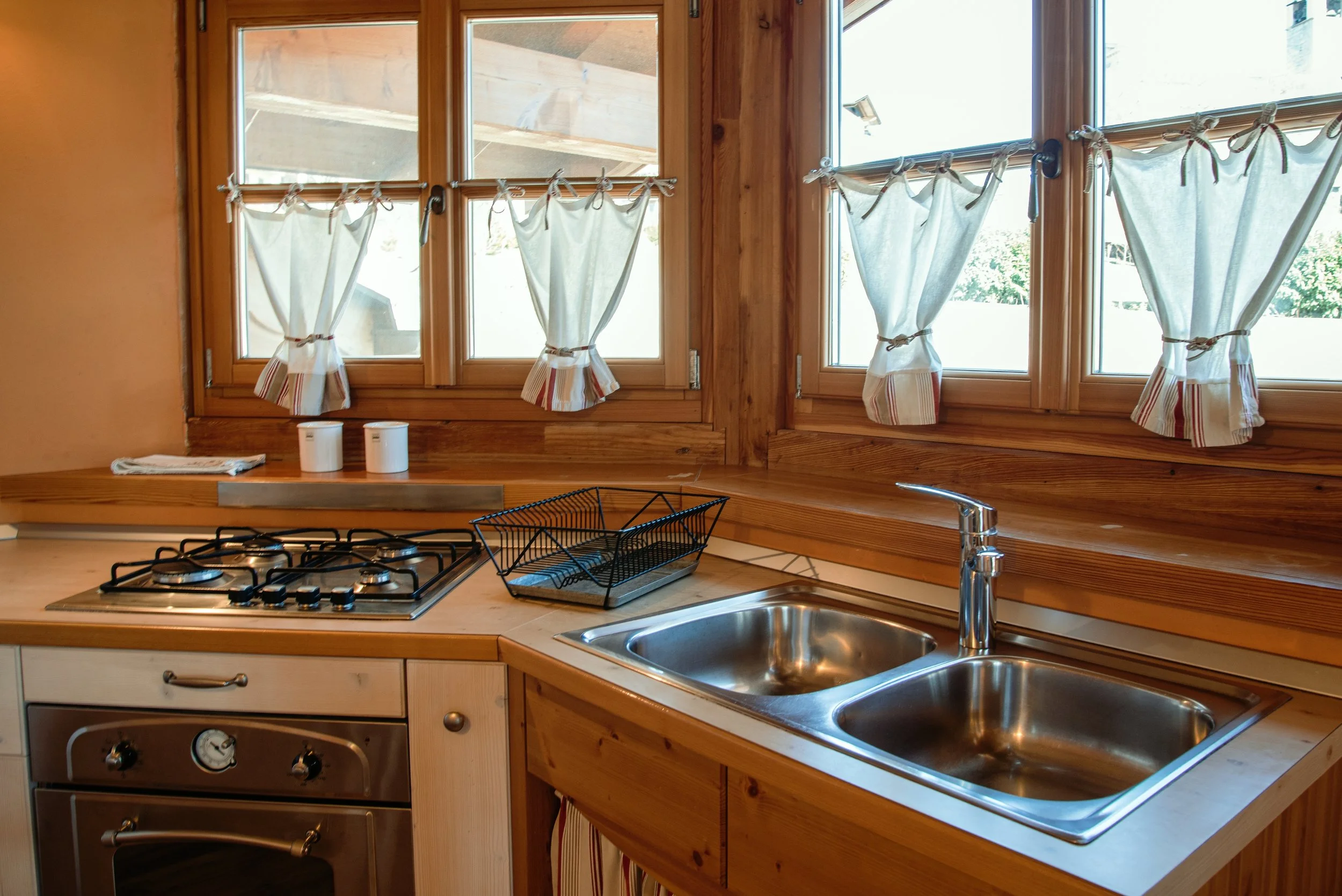 Cucina con lavello doppio in acciaio, piano cottura a quattro fuochi in metallo, finestre con tende di stoffa a righe e pannello di legno naturale.
