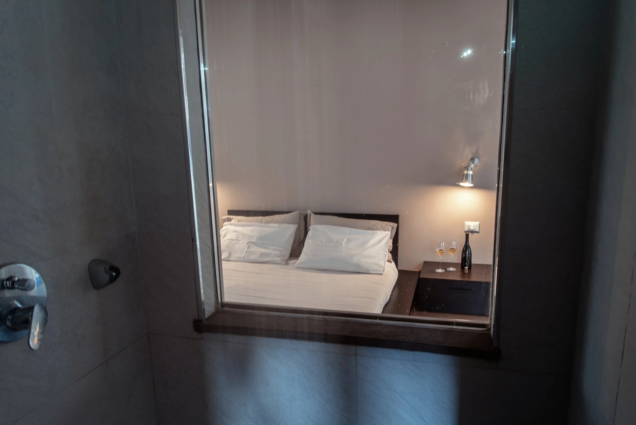 Specchio che riflette un letto con cuscini bianchi, una lampada da lettura e una mensola con due bicchieri di vino e una bottiglia nella camera da letto.