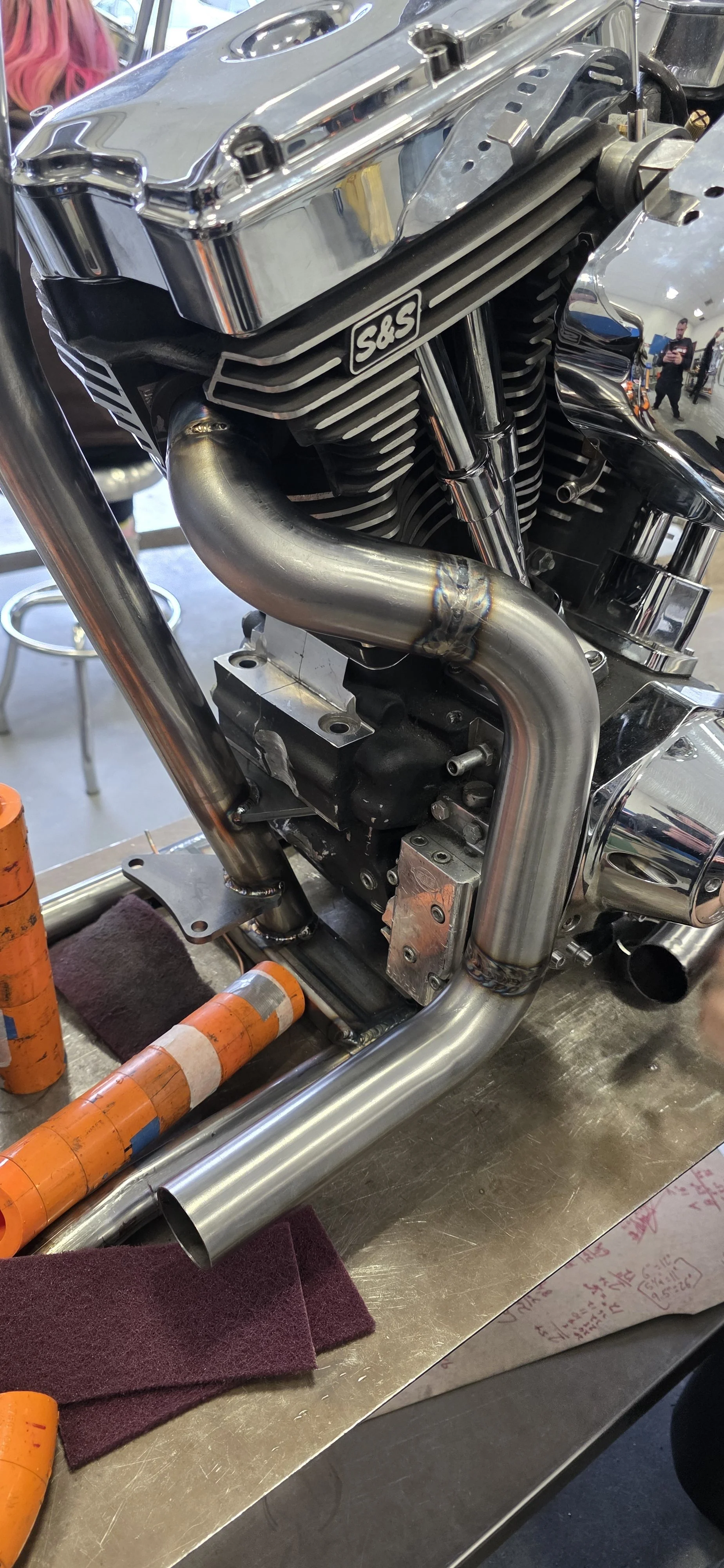 Close-up of a motorcycle engine with chrome and black components, featuring a prominent S&S logo. The engine has a custom exhaust pipe and is mounted on a metal workbench with tools and orange safety pins nearby.