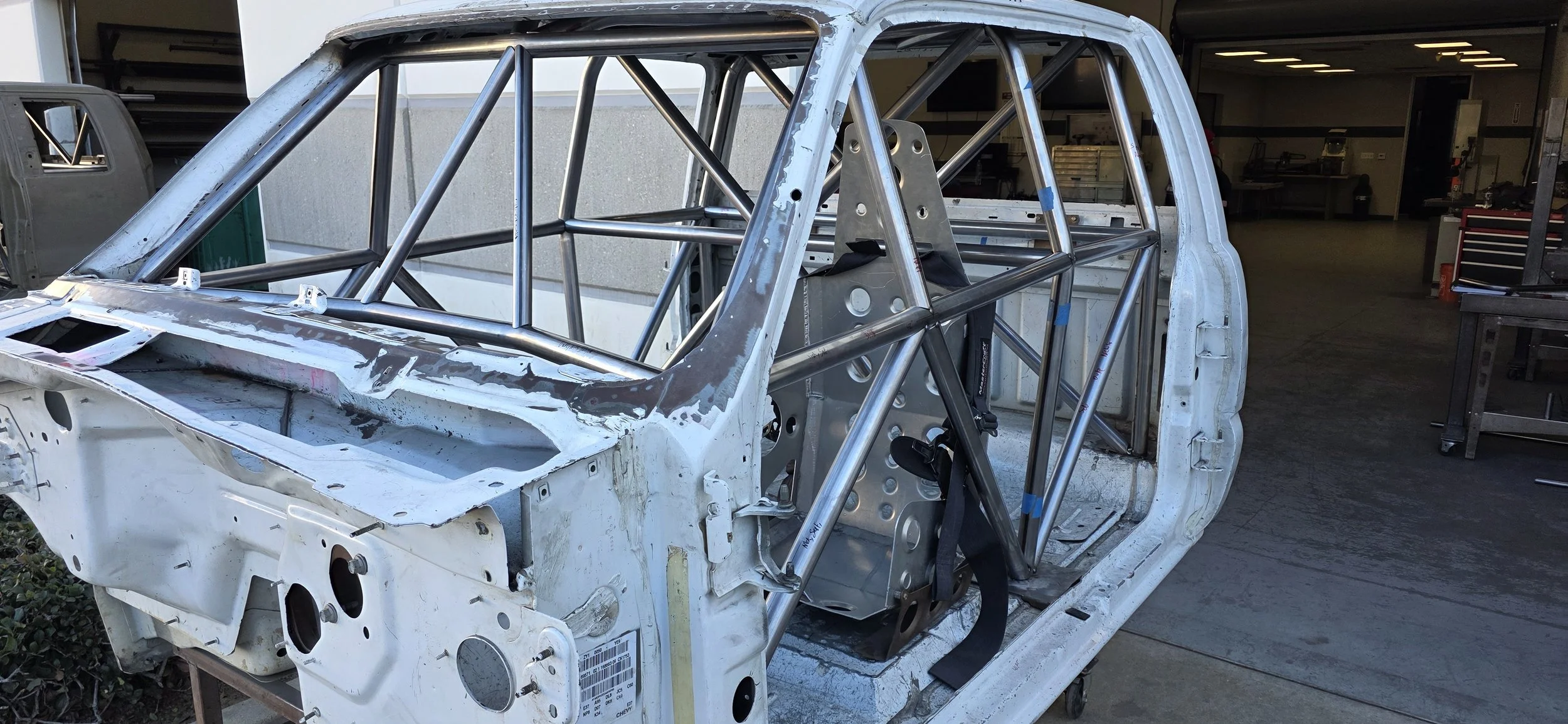 A partially assembled race car chassis with a roll cage inside, situated in a workshop or garage.