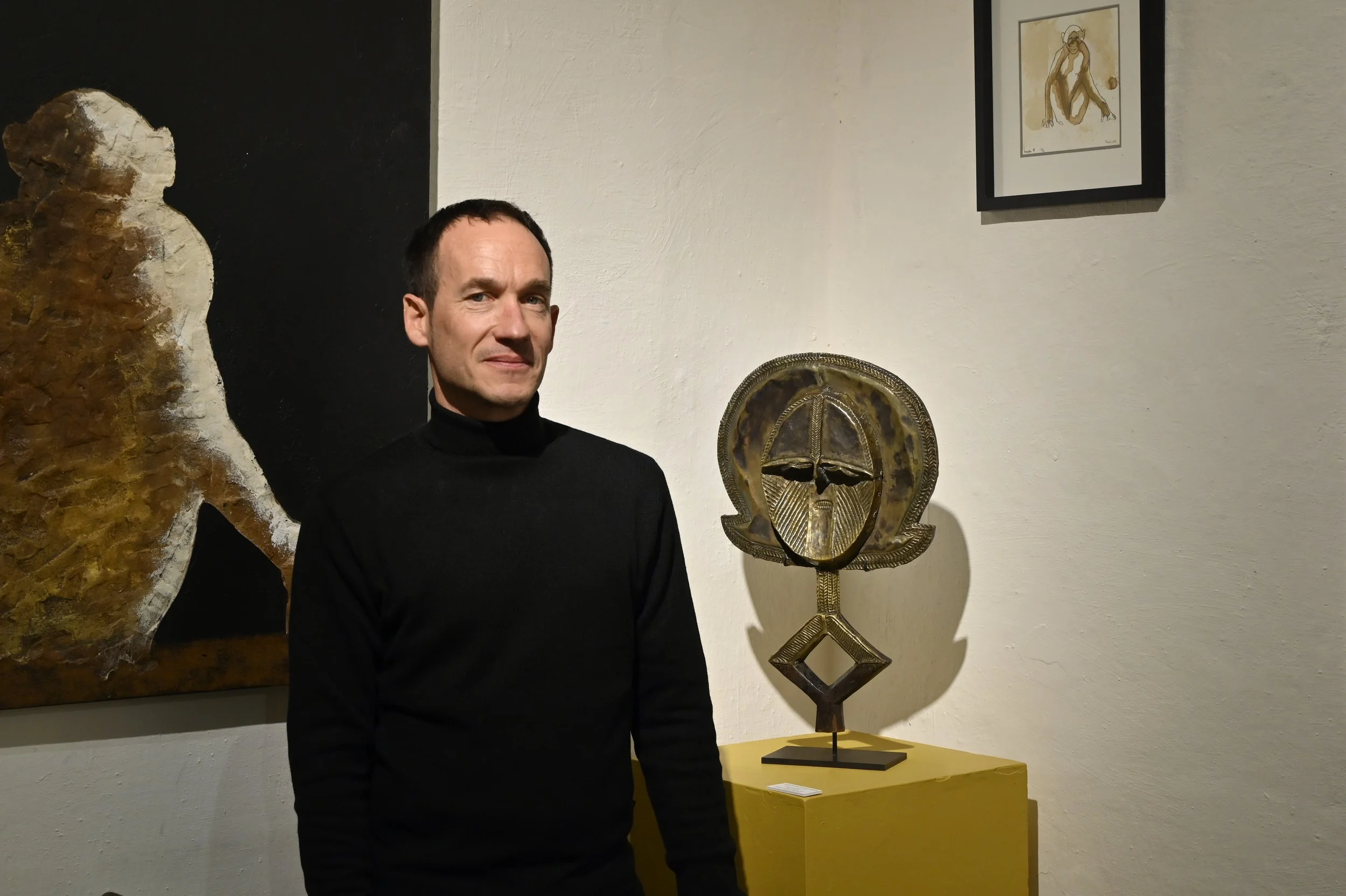Photo de Guénael Fassier,  debout dans la galerie Agama, portant un pull noir, à côté d'une sculpture en métal, reliquaire Bakota du Gabon reposant sur une colonne jaune, avec des peintures de l'artiste Pavan en arrière-plan.