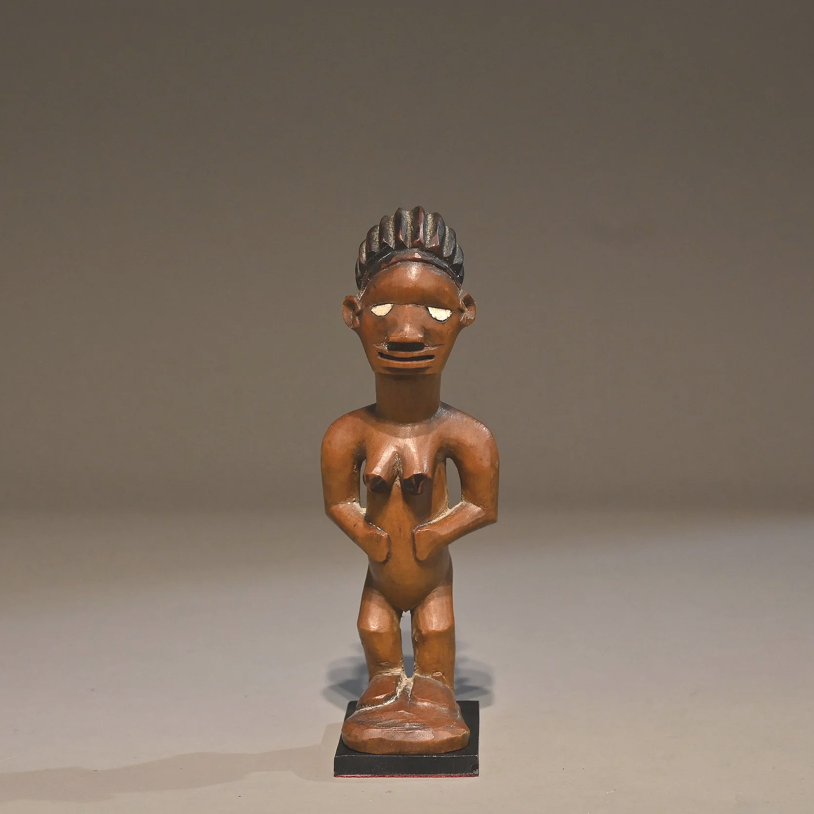 Statue en bois, des Bembe du Congo,  représentant une figure humaine stylisée avec des cheveux en relief, sans vêtements, posant avec les mains sur le ventre, sur une base noire.