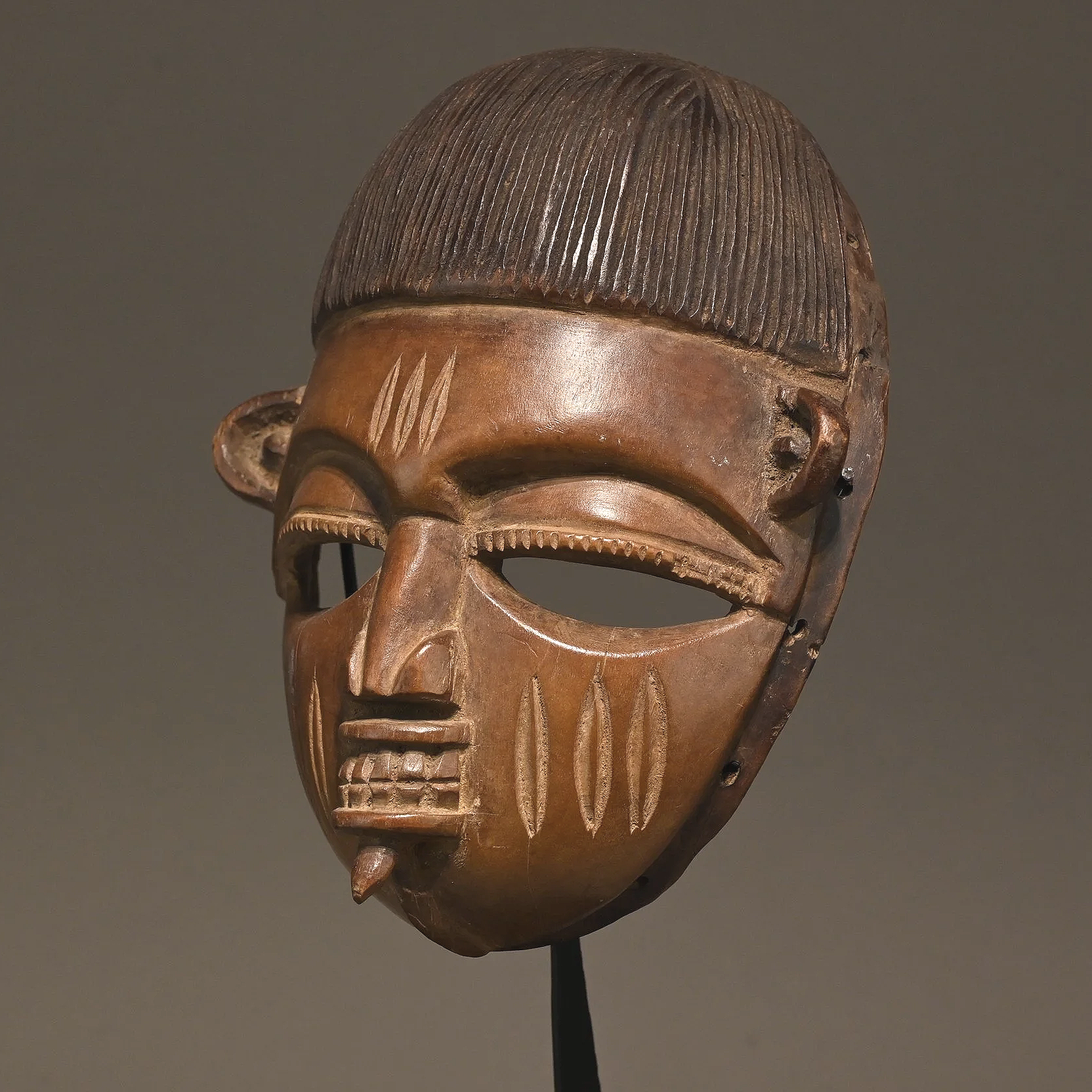 Masque de danse Egun du groupe culturel yoruba (Nigéria) représentant un visage circulaire de couleur marron miel, les yeux creux en amandes, la bouche ouverte montrant les dents, un labret dans la lèvre inférieure. La coiffe est légèrement stylisée.