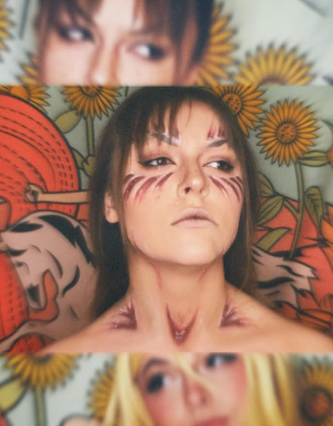 A person with face paint resembling tiger stripes and markings on their face, neck, and shoulders, lying on a background with sunflower print and illustrated tiger designs.