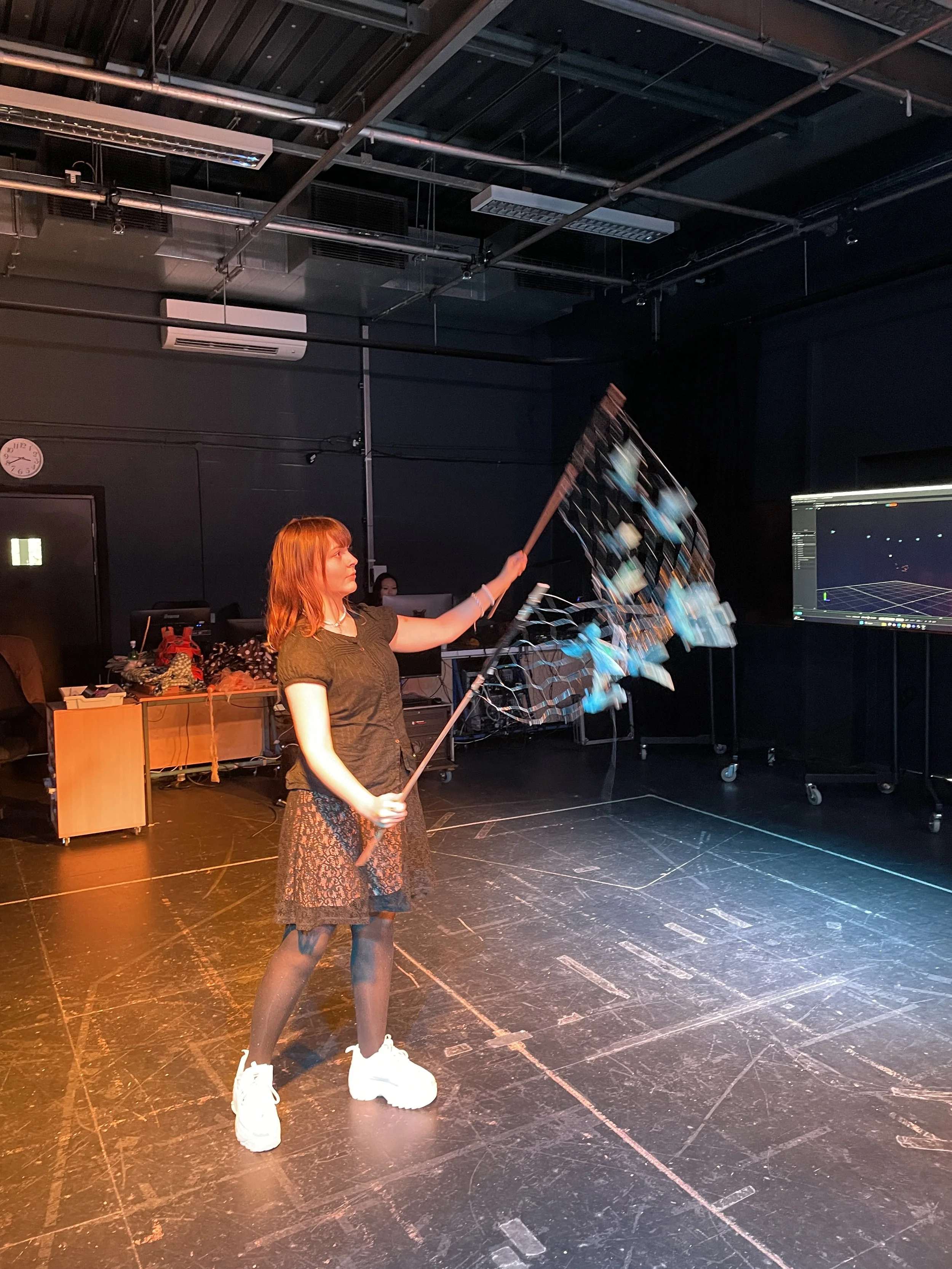 A young woman with red hair, dressed in a black top, a patterned skirt, sheer tights, and white platform sneakers, is standing on a dark stage. She is holding a long stick with multiple blue and white paper fans attached, creating a visual effect as 