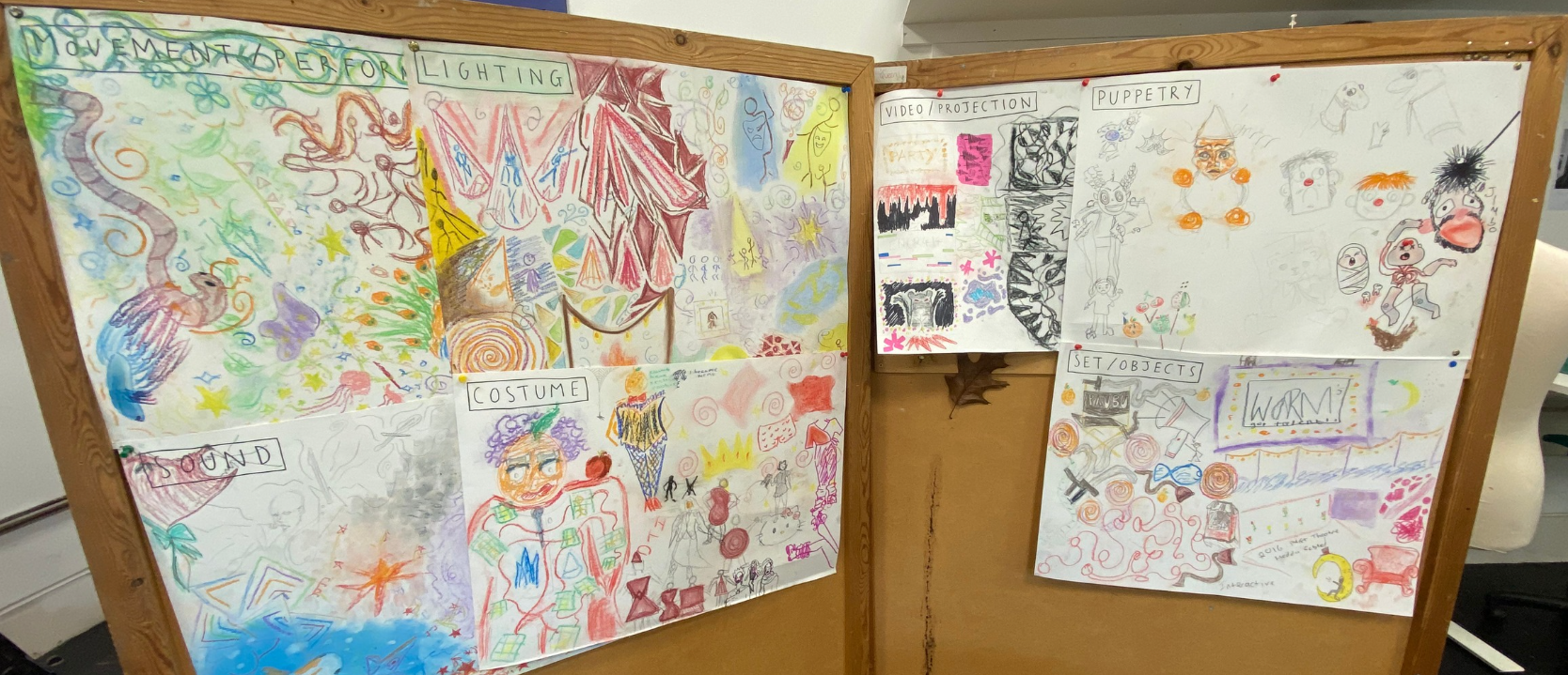 A wooden display board with colorful, hand-drawn posters related to a performance or production. The posters include sections labeled 'MOVEMENT & PERFORMANCE,' 'LIGHTING,' 'VIDEO/PROJECTION,' 'PUPPERY,' 'COSTUME,' 'SET/OBJECTS,' and 'WORM!'. The draw