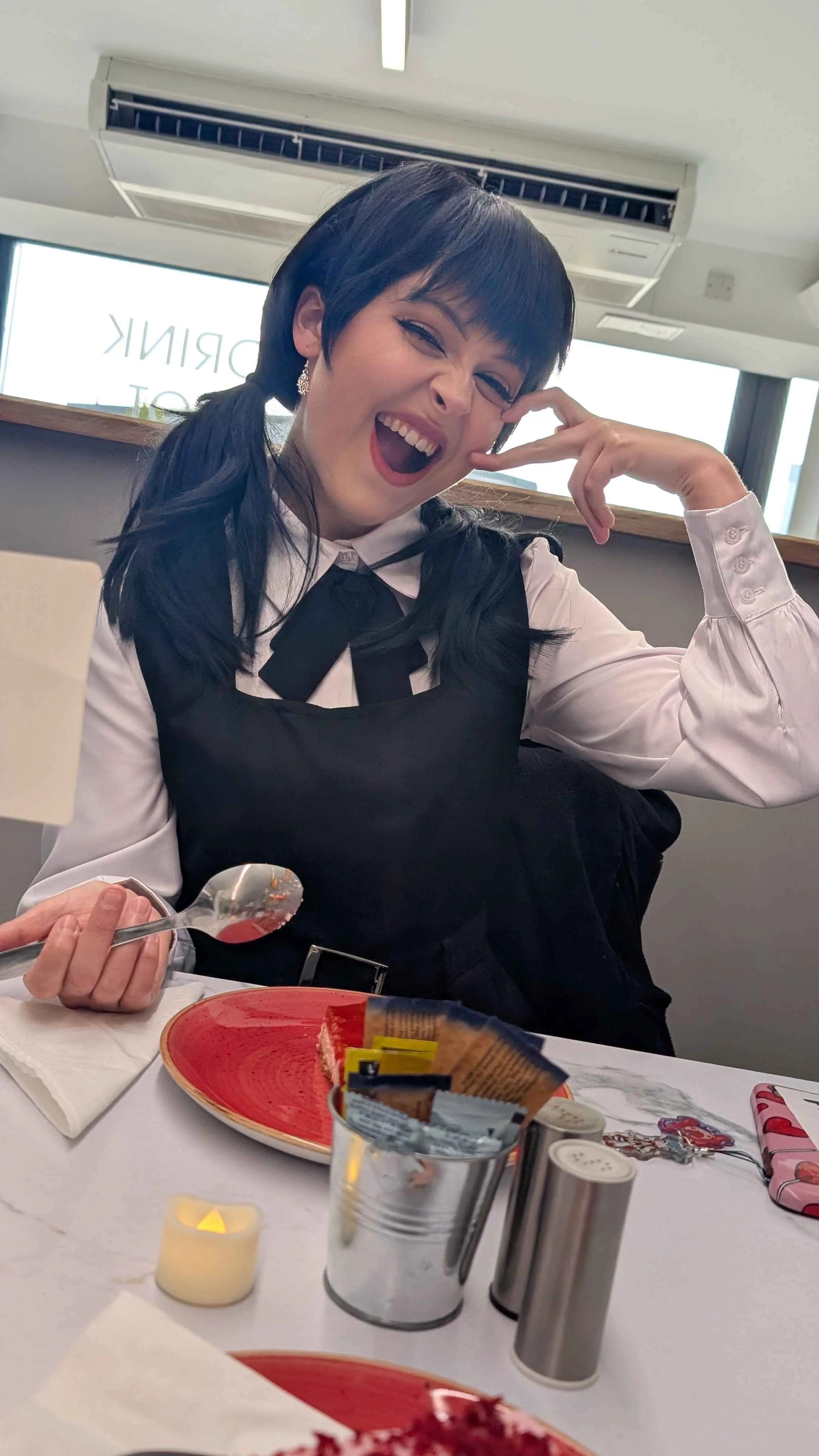 A woman with short black hair wearing a white blouse and black vest, smiling and making a playful gesture with her hand near her face, sitting at a table with a red plate, a spoon, and various condiment containers, in a bright indoor setting with lar