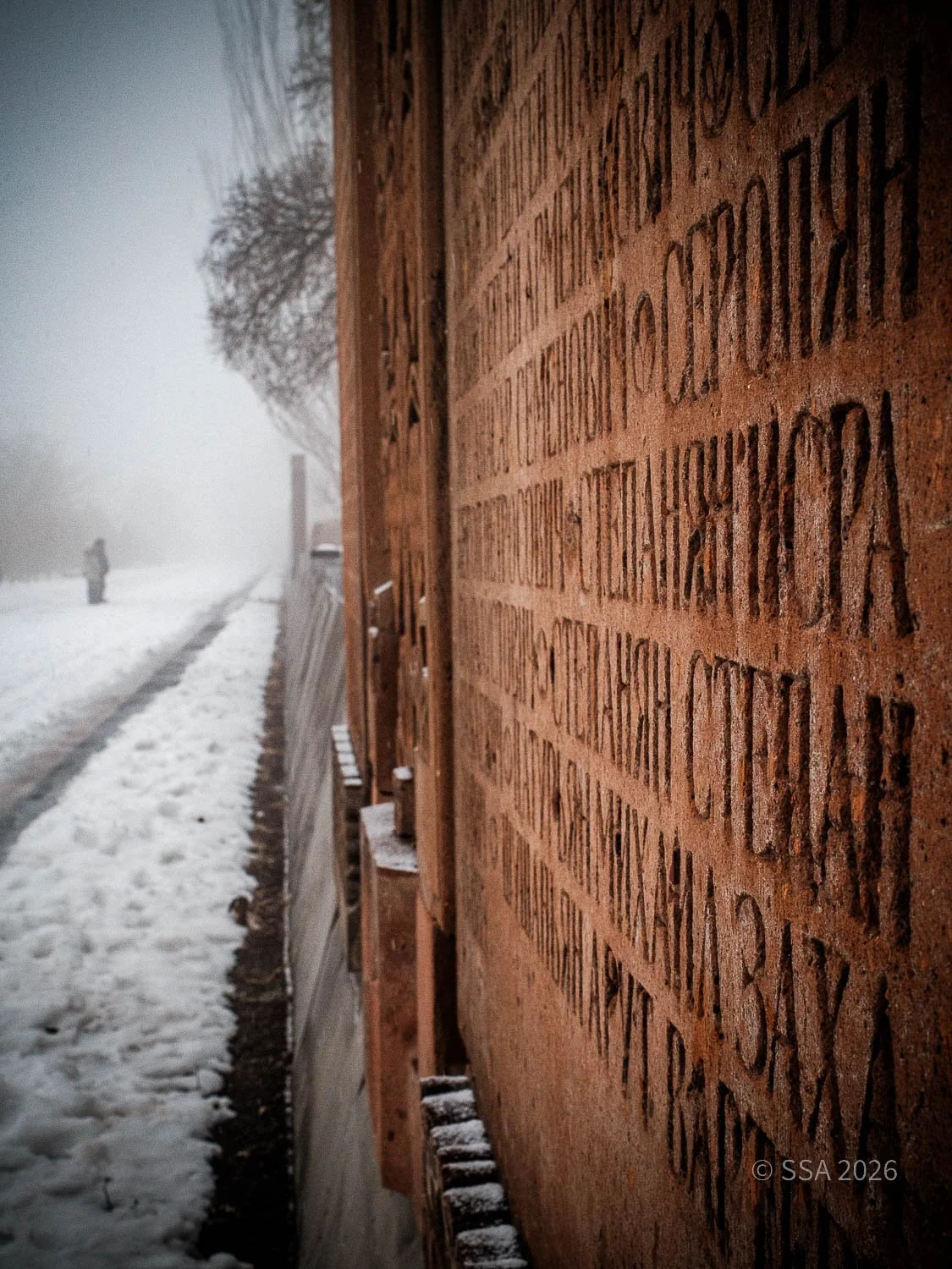 Close-up of a brick memorial wall with engraved names, snow on the ground, and a person walking in the misty background.