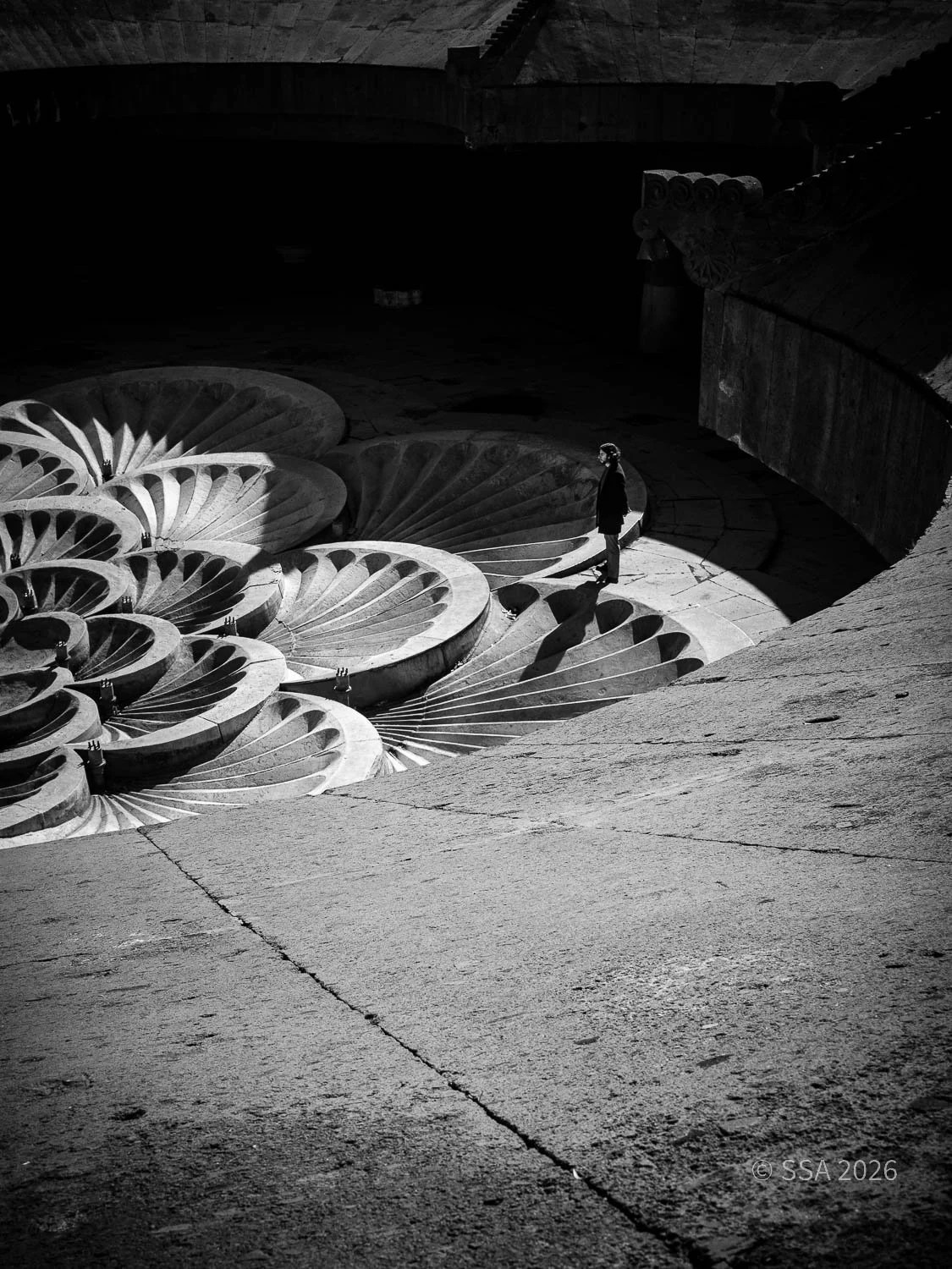 A person standing alone on a curved stone pathway with large shell-shaped structures beneath in a dark, shadowed setting.