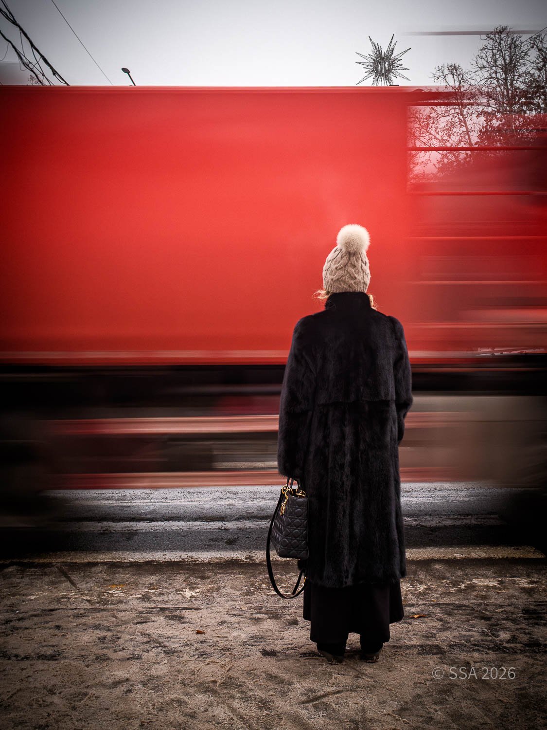 Person standing on a sidewalk looking at a moving red truck, wearing a black coat, cream knit hat with pom-pom, holding a black handbag.