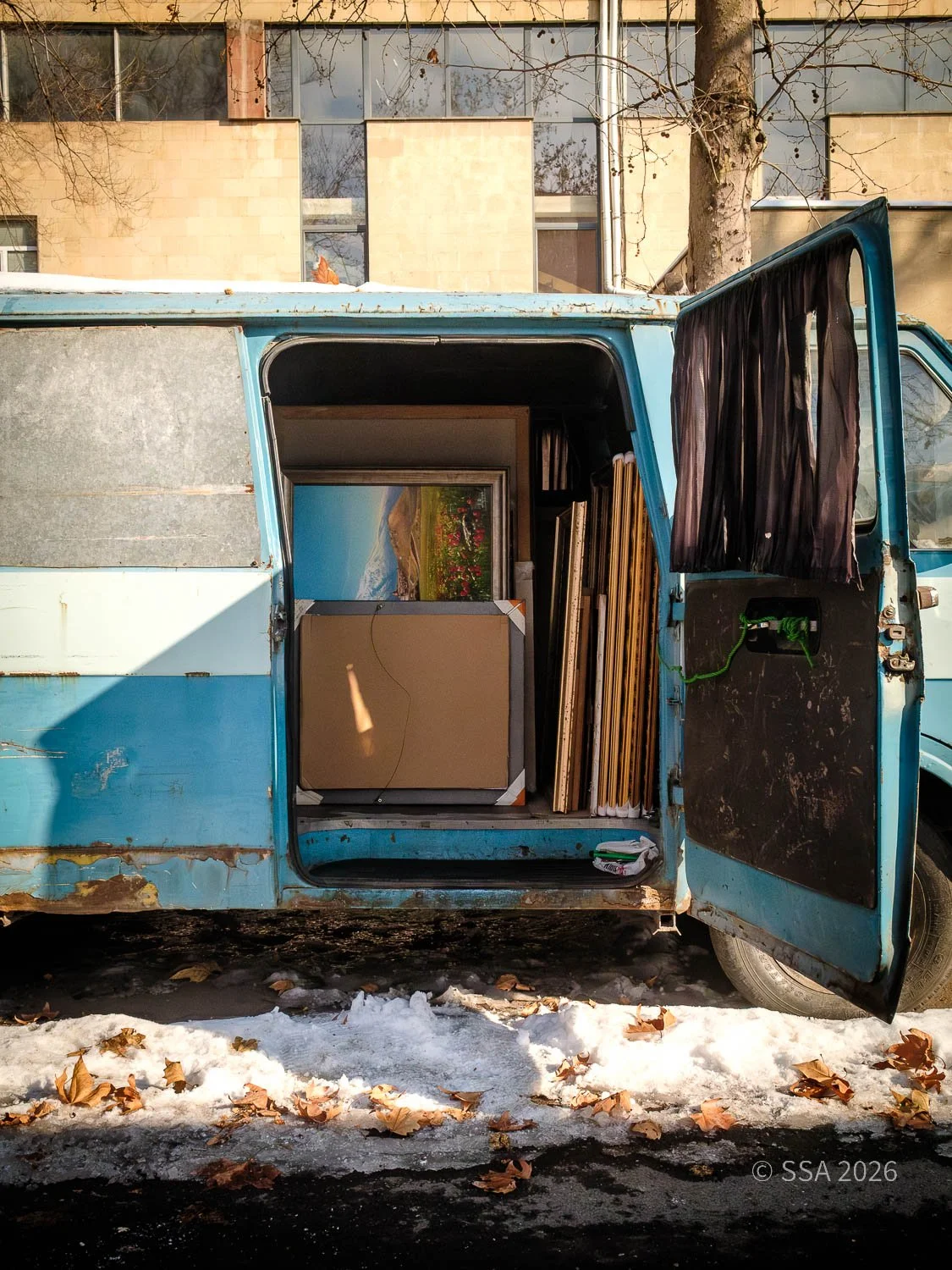 Open blue truck filled with framed paintings and art prints, parked outdoors on a snowy street in front of a building with large windows.