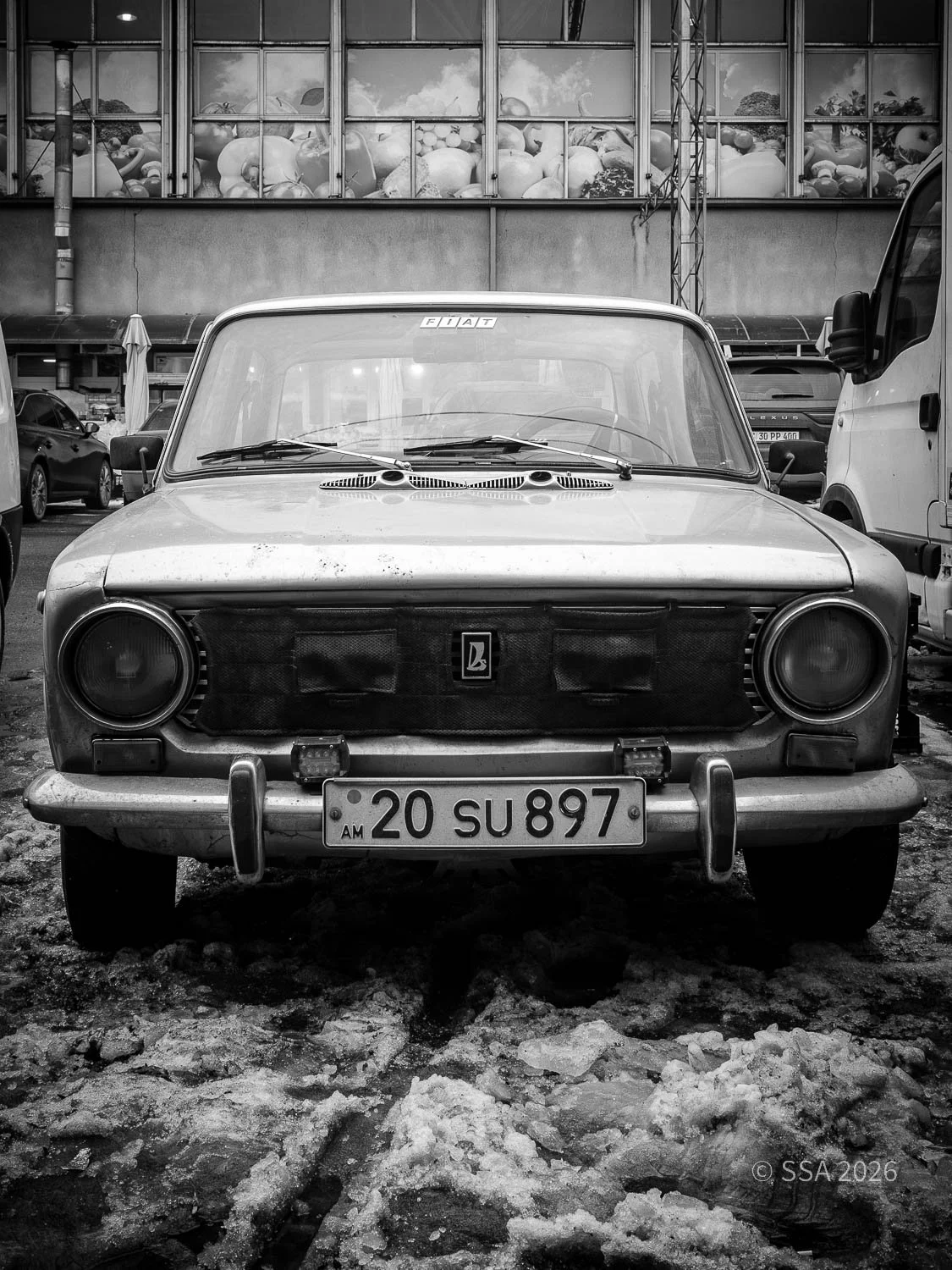 Old Fiat car parked in a lot with snow and ice on the ground. The car is weathered, with a black cover over the front grille and a license plate reading '20 SU 897'. There are other vehicles and a building with large windows in the background.
