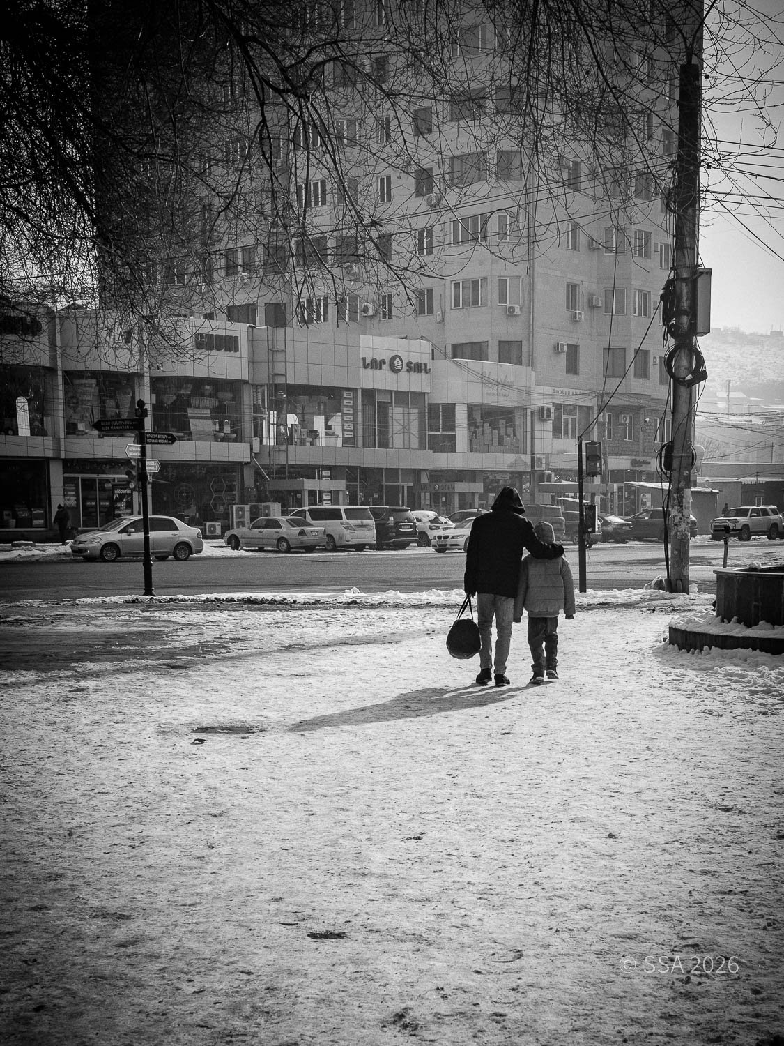A black-and-white photo of a person and a child walking on a snowy sidewalk in an urban area with cars parked along the street. The person has their arm around the child, and the person is carrying a bag. The background shows a large building with mu