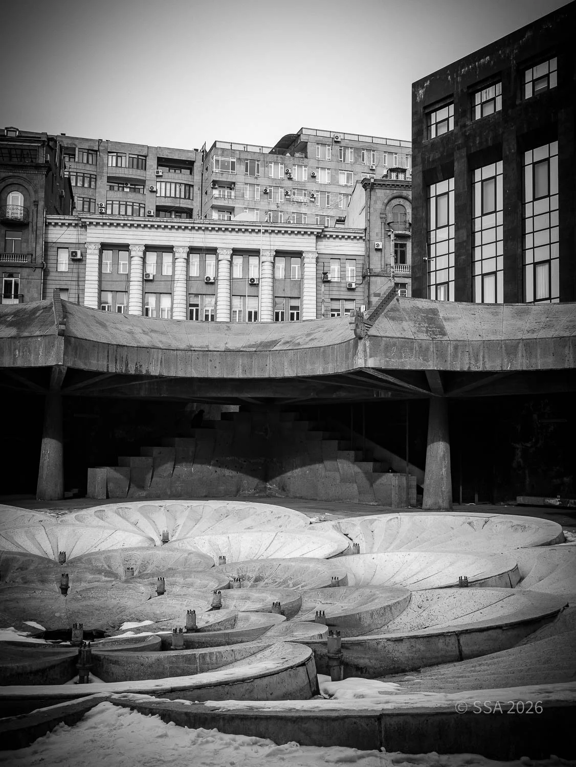 Black and white photo of an outdoor urban scene with a multi-level concrete structure and large flower-shaped basins in the foreground, surrounded by tall buildings.