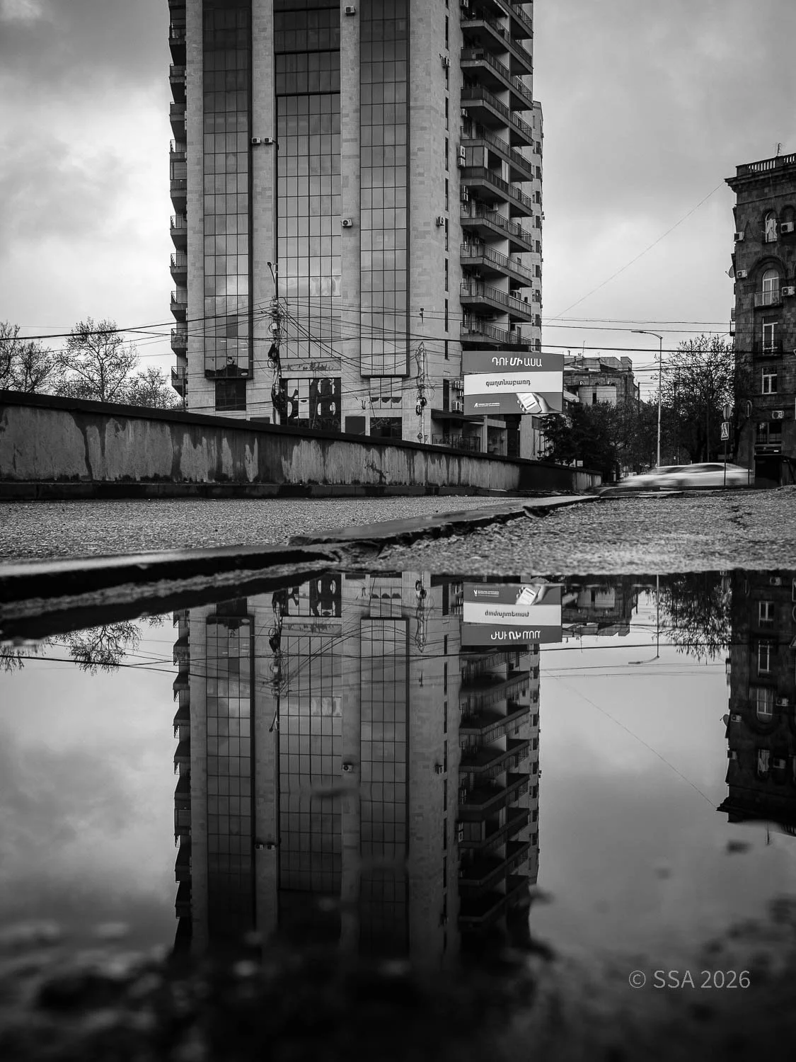 Black and white photograph of a tall building reflected in a large puddle on the street, with the cloudy sky overhead.