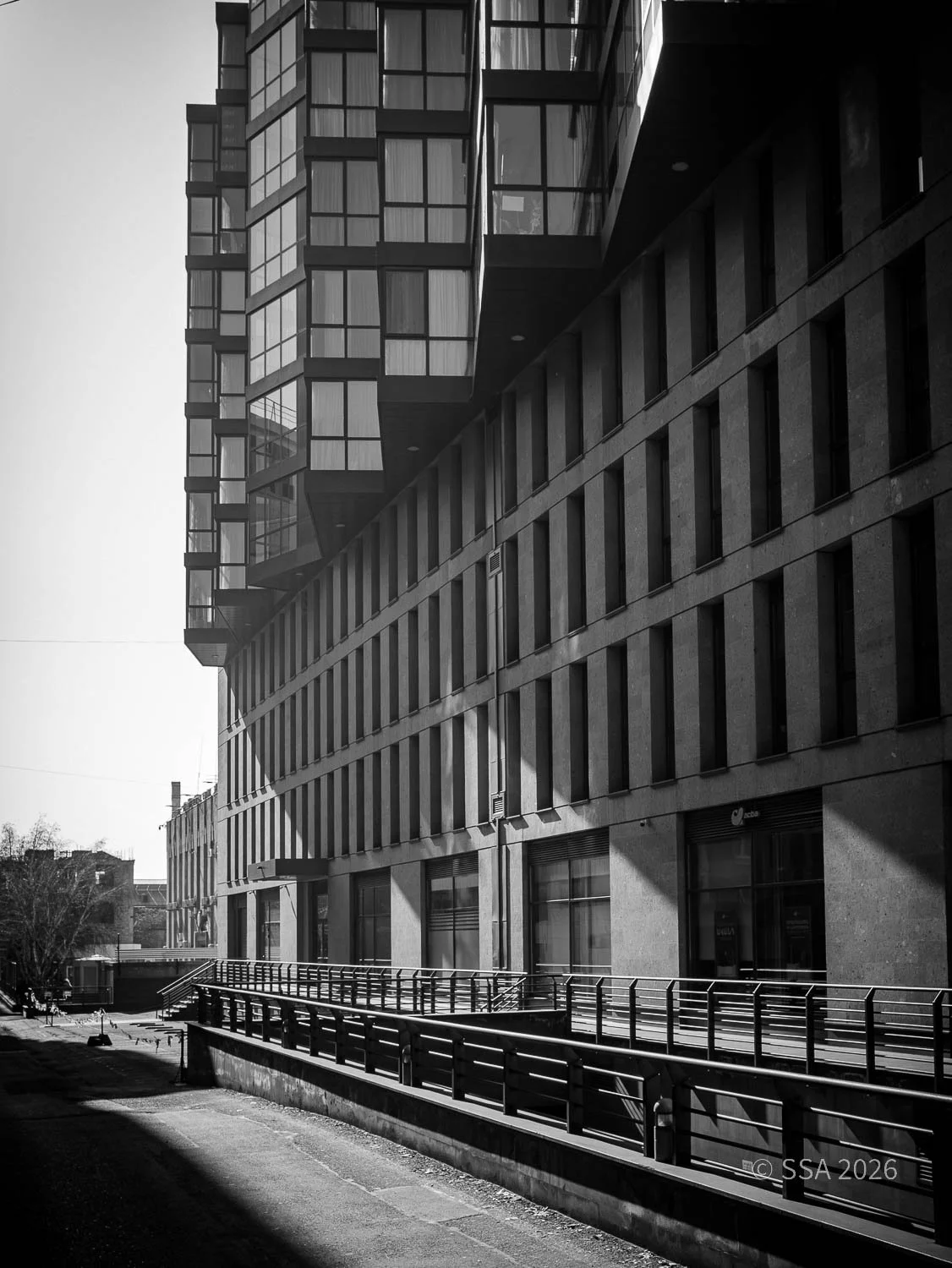 Black and white photo of a modern multi-story building with balconies and large windows, with a urban street and sidewalk in the foreground.