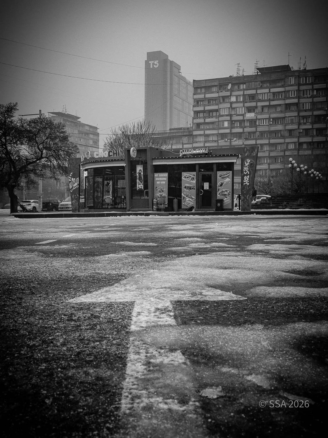 Black and white photo of a small fast food stand in an urban area, with high-rise buildings in the background. The photo is taken from a low angle, showing an arrow painted on the icy street pointing towards the stand.