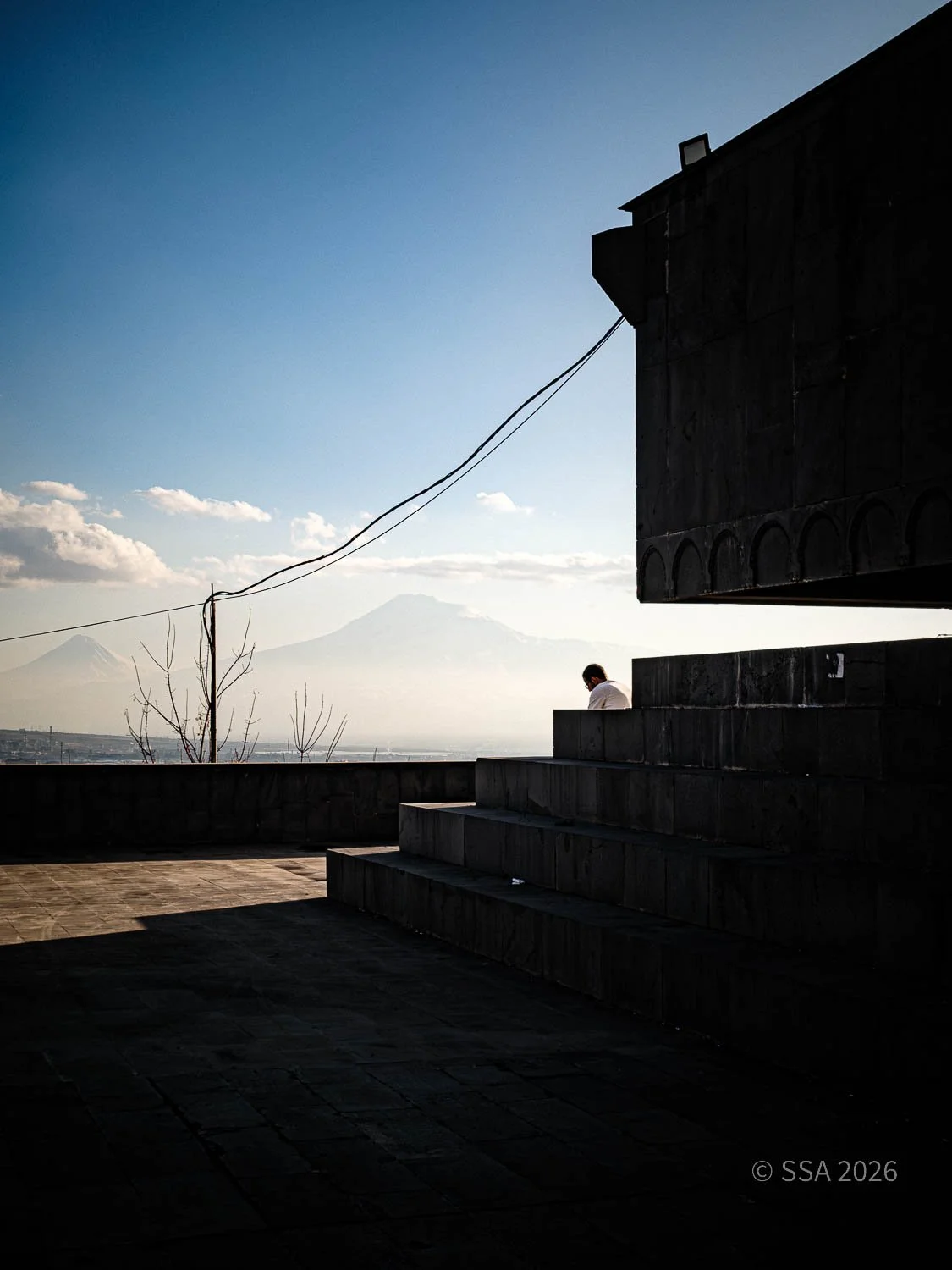 A silhouetted person sitting on stairs outside in the late afternoon or early evening with mountains in the background, including Mount Fuji, and a clear sky with some clouds.