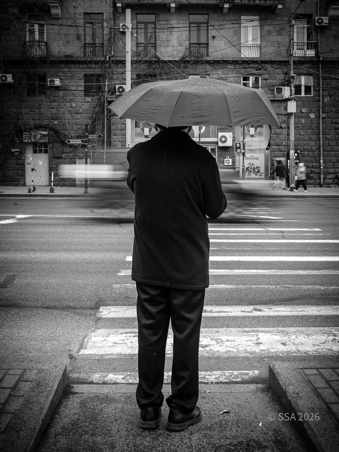 A person in dark clothing standing at a crosswalk on a rainy day, holding an umbrella, with a blurred background of city buildings and passing cars.