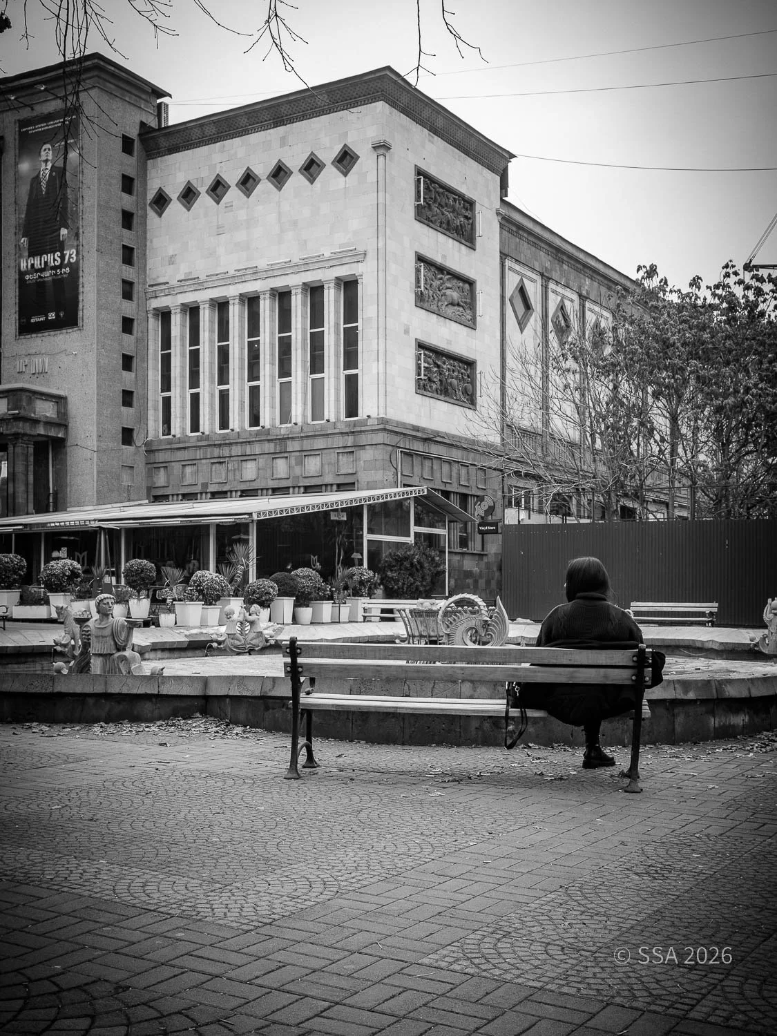 A woman sits on a park bench facing a decorative fountain in an urban park, with large buildings in the background and trees with bare branches overhead. The scene is in black and white.