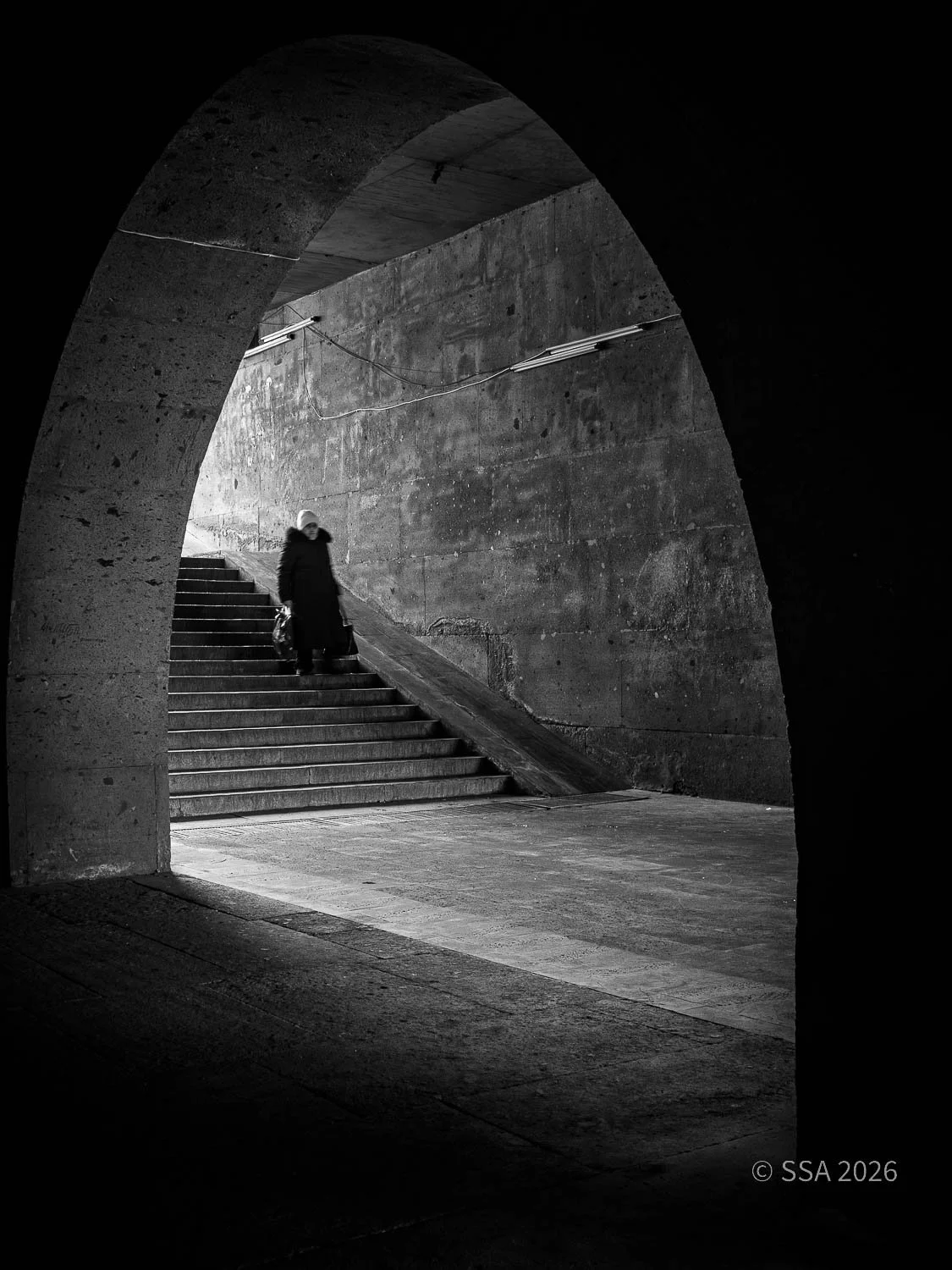 Black and white photo of a person descending stairs in an underground tunnel or passage, viewed through an archway.