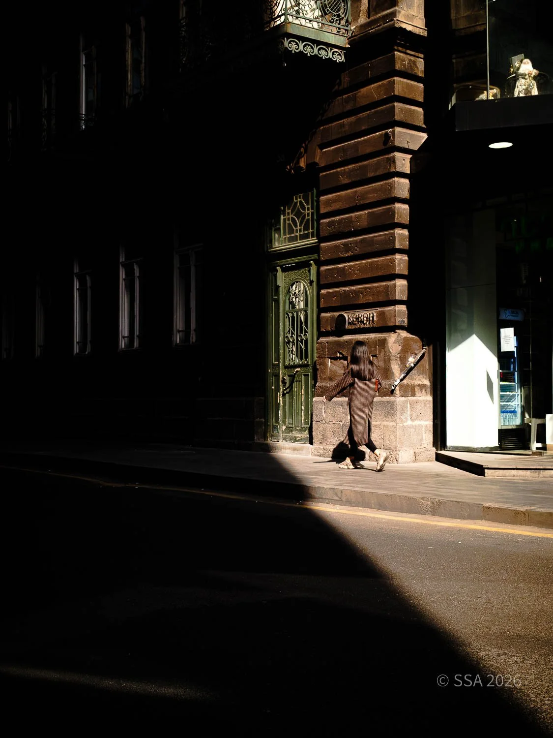 A person with long hair and dark clothing walking past an old building with green doors and ornate ironwork in a city street, with a sharp shadow cast on the sidewalk.