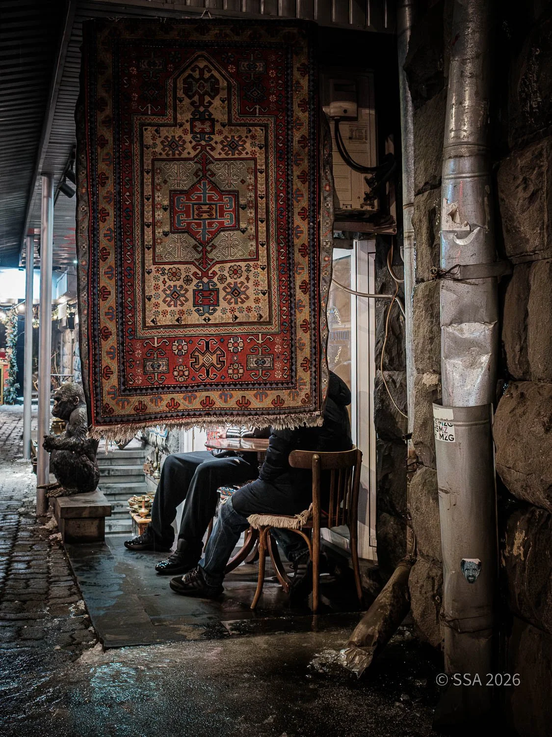 A small shop with a colorful, patterned rug hanging at the entrance. Inside, two people are sitting at a small table, partially obscured by the rug. There is a stone wall on one side and a cobblestone street outside. Various objects and sculptures ar