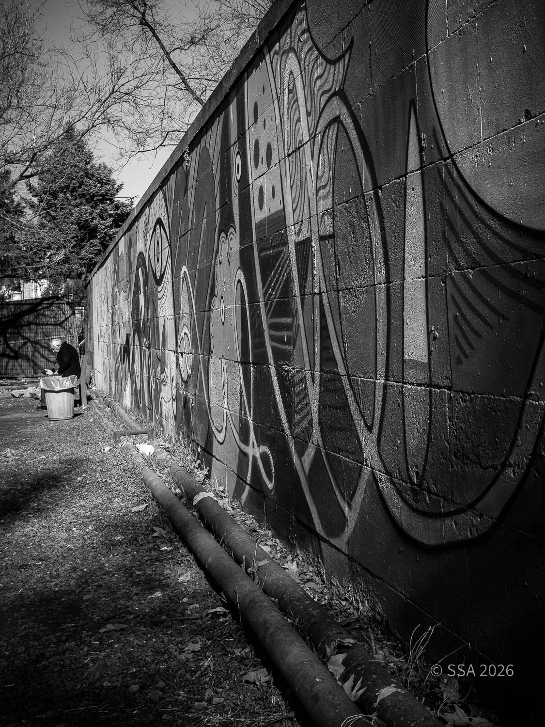 Black and white photograph of a graffiti mural on a concrete wall, with two pipes at the bottom and a person standing near a trash can in the distance.