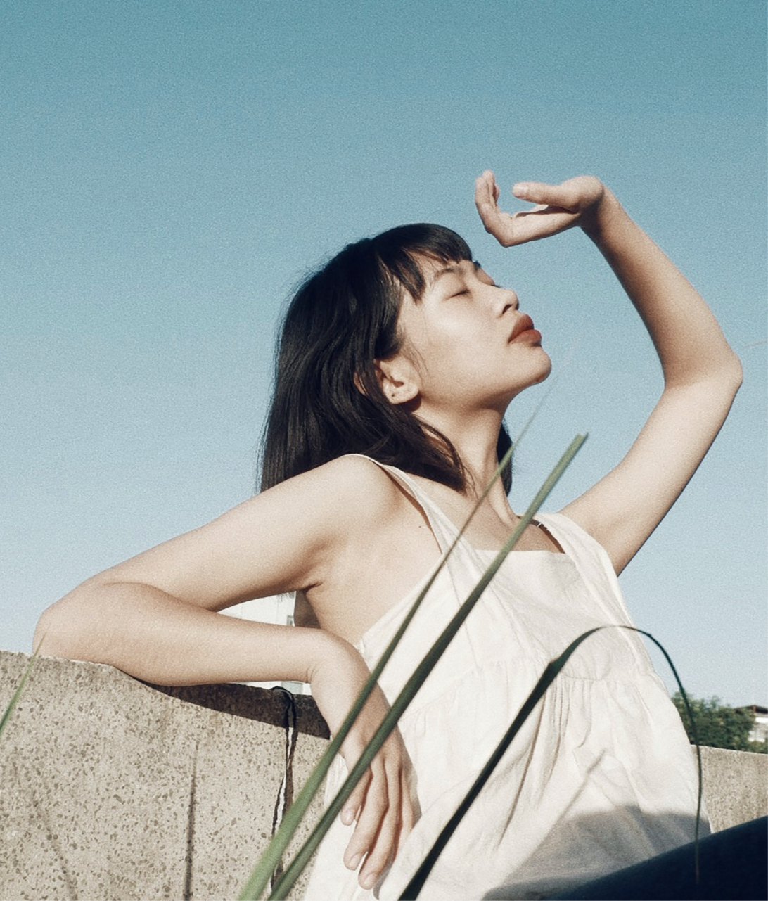 A woman with dark hair and bangs is outdoors, sitting and leaning on a stone surface, with her eyes closed and a peaceful expression. She has one arm resting on the stone and the other hand raised to her forehead, shadowed by the sunlight. The background shows a clear blue sky and some distant trees.