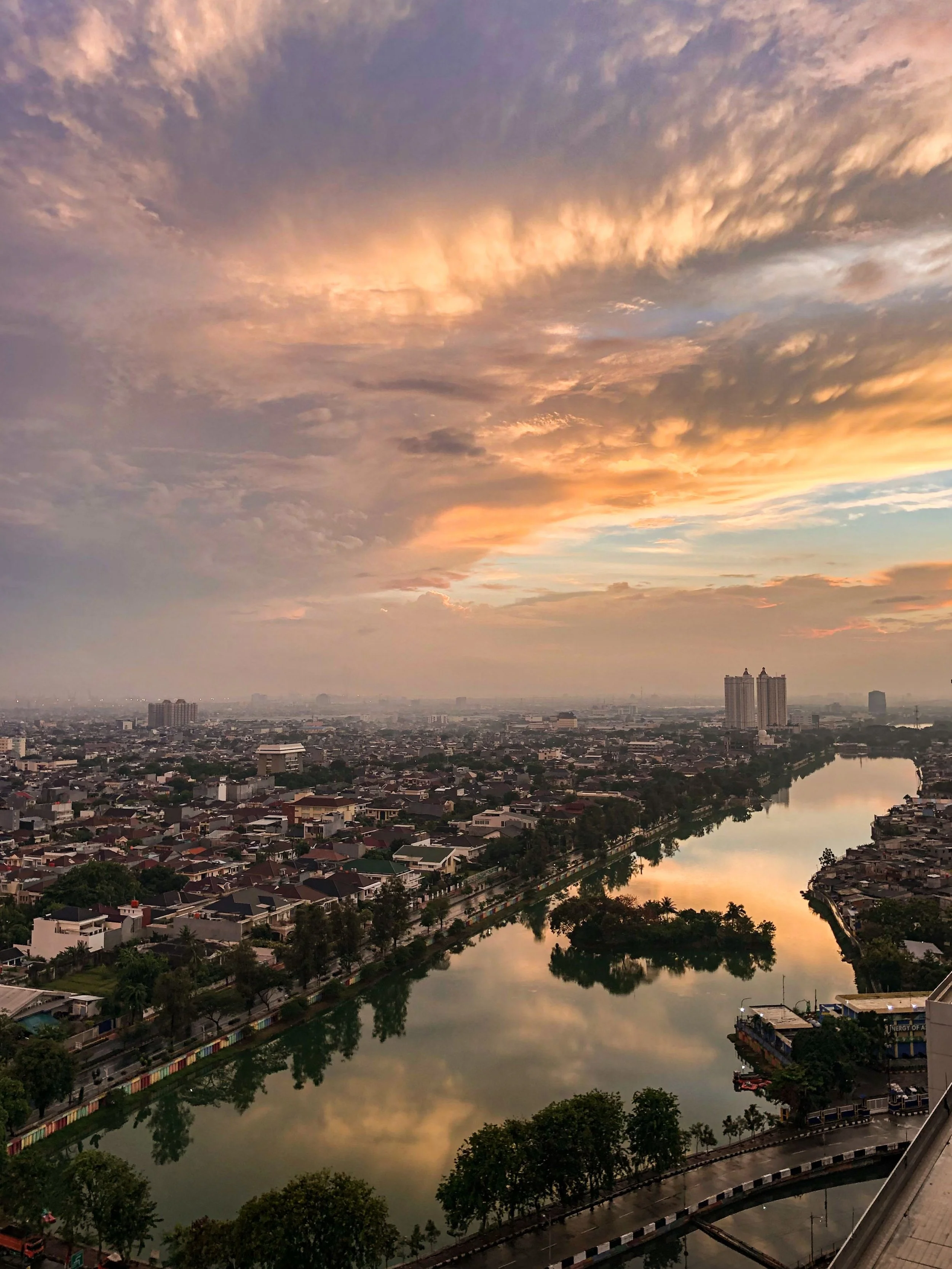 Cityscape during sunset with a river reflecting the colorful sky and a mix of high-rise and residential buildings.