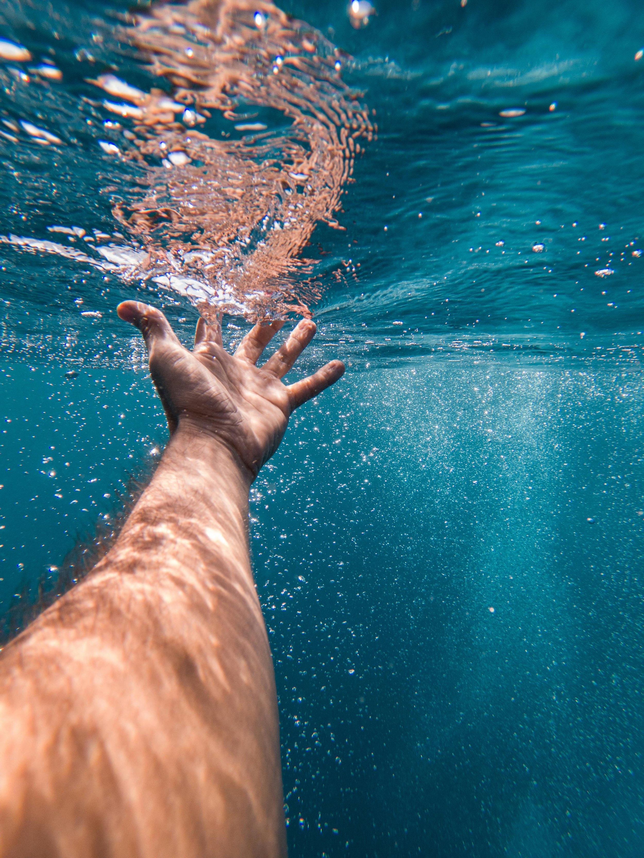 A person's arm reaching upward underwater, with bubbles and ripples on the water surface.